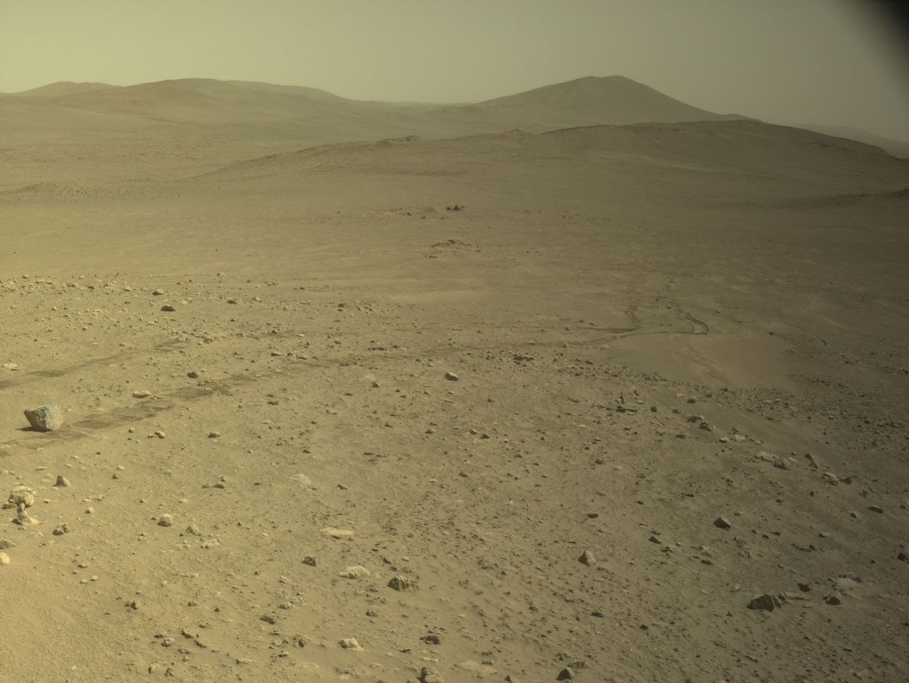 航天图片资料 · A wide view of the Martian surface taken by the Perseverance rover. The foreground features an arid, tan-colored terrain scattered with rocks and pebbles of varying sizes. Faint, parallel rover tracks curve through the dirt from the middle-left towards a shallow sandy depression in the midground. A distinct, slightly larger rock sits near the tracks on the far left. The background consists of smooth, gently rolling hills and distant ridges that fade into a hazy, pale yellow-brown sky.