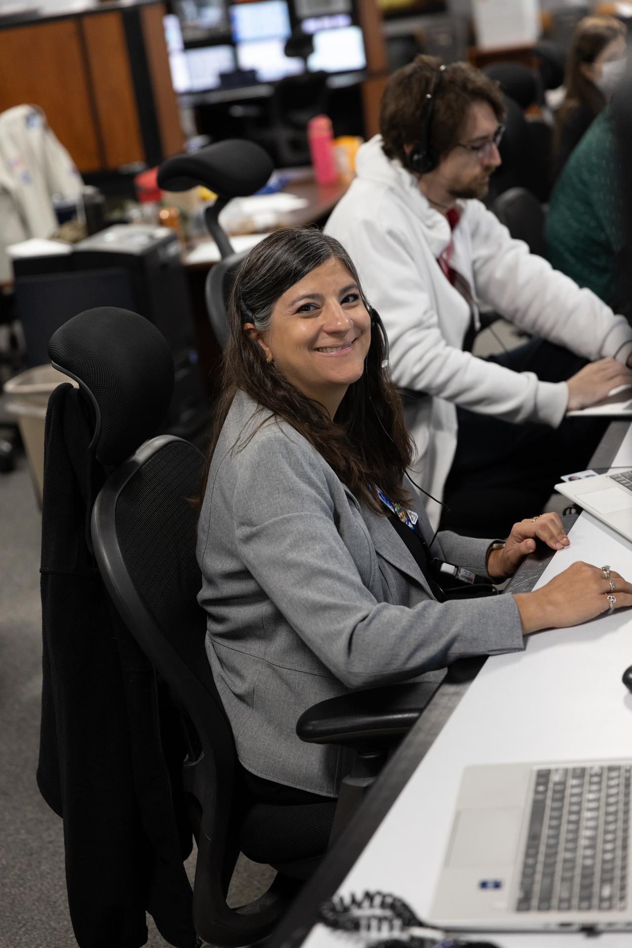 航天图片资料 · A women with dark brown hair smiles at the camera for a photograph. The woman is wearing a grey jacket and a headset while she sits at the desk with a computer;
