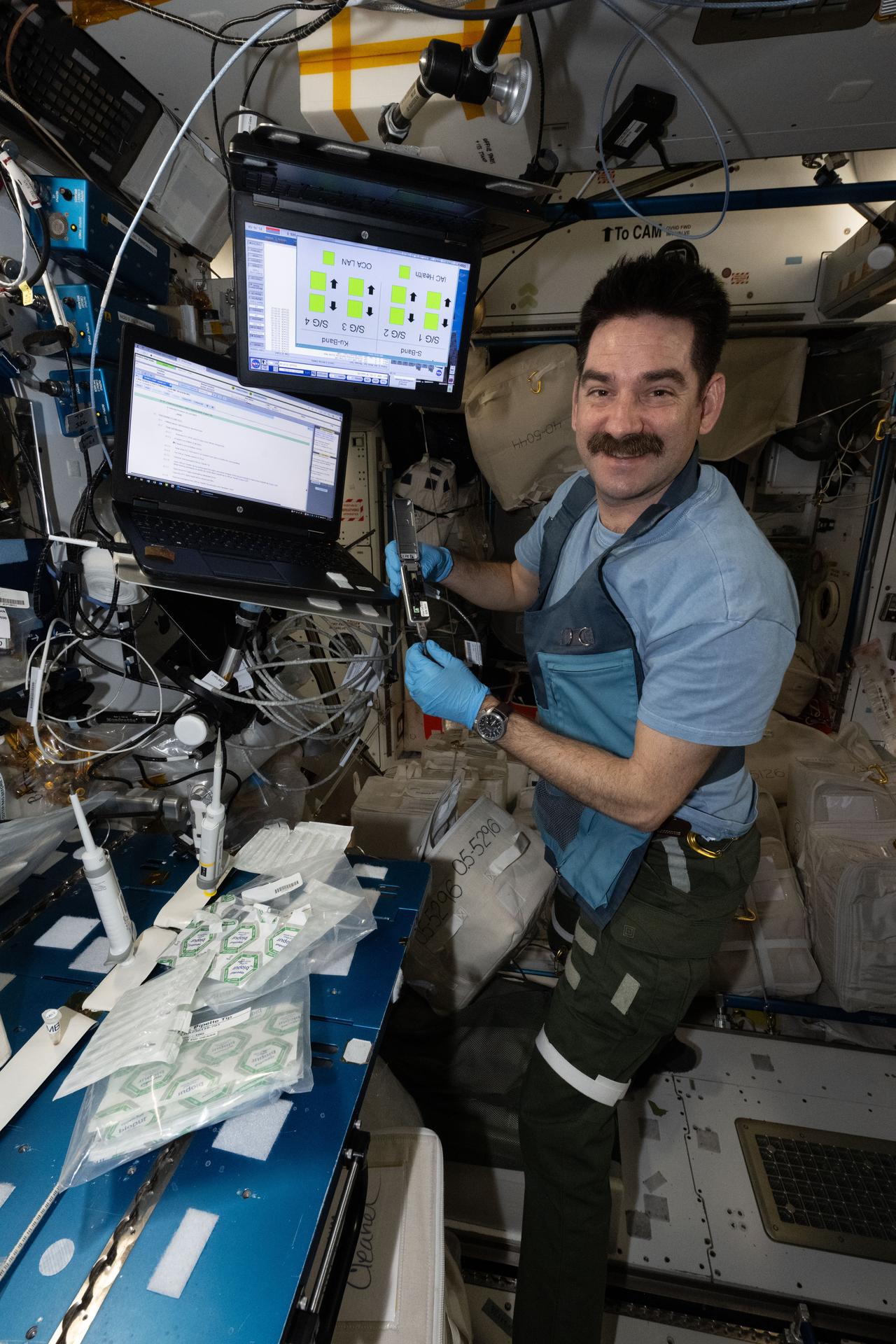 航天图片资料 · NASA astronaut Jack Hathaway works aboard the International Space Station. He is wearing blue gloves while holding a metallic device with a hinge at its center. To his left are several plastic packages and a pipette, with two laptop screens mounted above the work area.