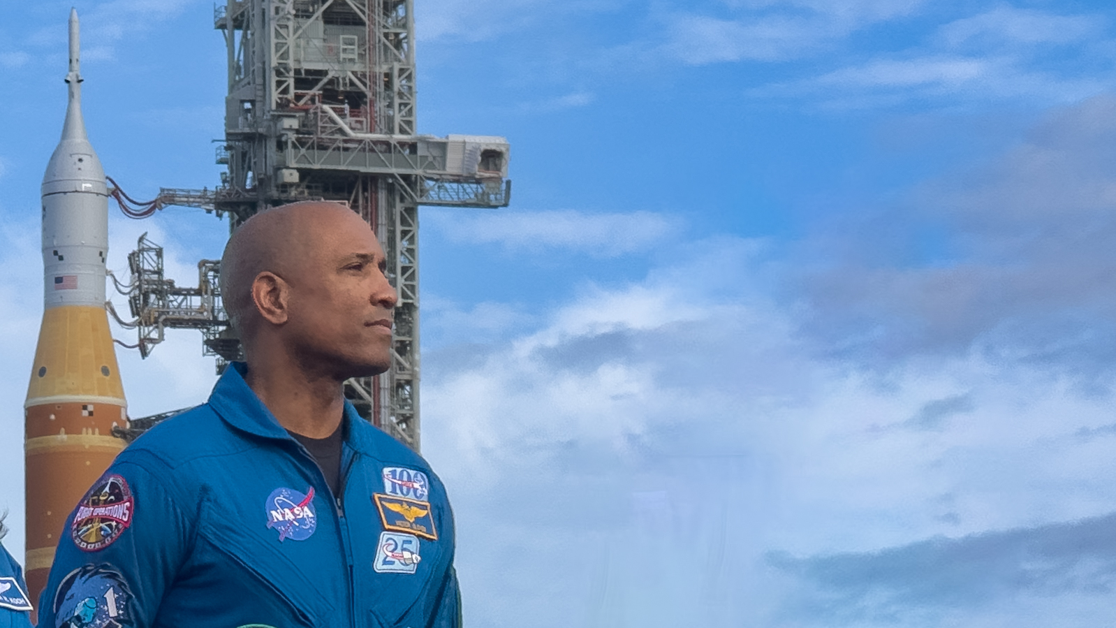 航天图片资料 · A man in a blue jumpsuit stands against a blue sky with a big orange rocket behind him on the left.
