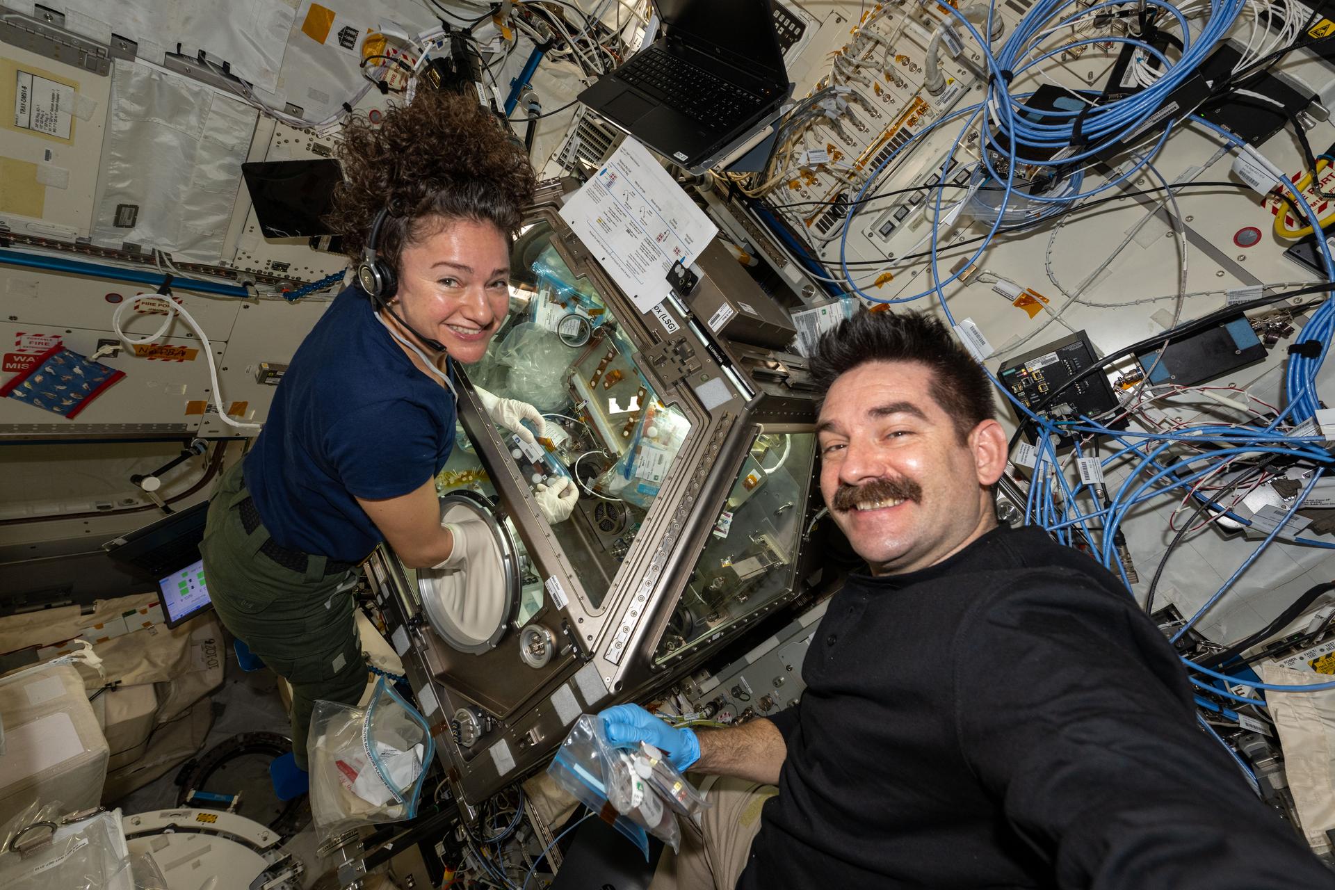 航天图片资料 · NASA astronauts Jessica Meir (left) and Jack Hathaway (right) smile at the camera as Hathaway takes a selfie aboard the International Space Station. Meir wears blue gloves while performing operations inside the Life Sciences Glovebox, surrounded by metallic panels, cables, and equipment.