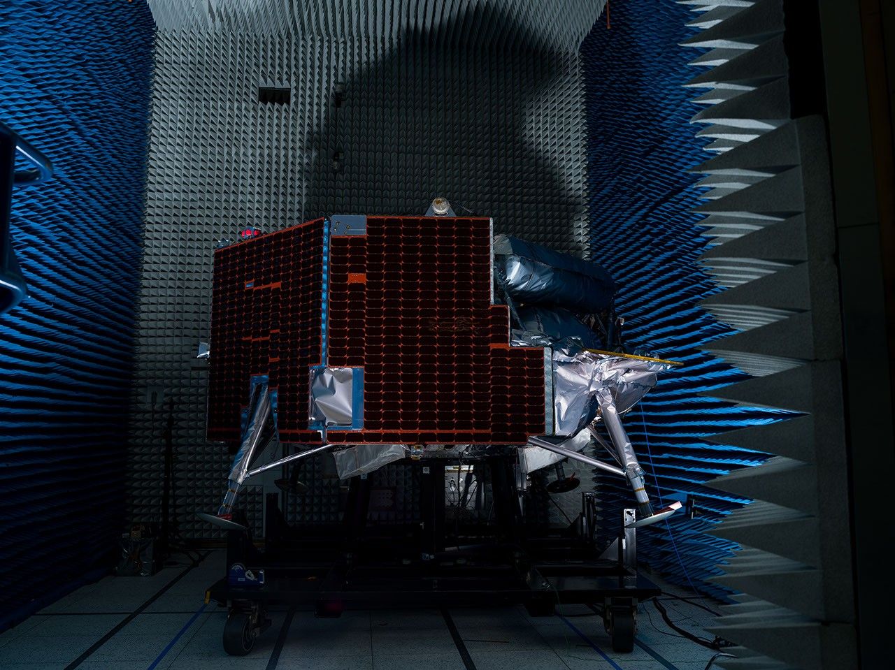 航天图片资料 · A lunar lander equipped with dark solar panels and foil-wrapped legs sits on a platform inside a dark anechoic chamber lined with blue and gray foam spikes.