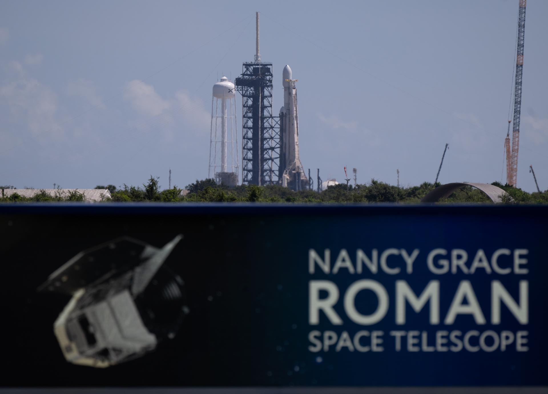 航天图片资料 · Image shows a banner that reads Nancy Grace Roman Space Telescope. In the background is a rocket standing vertically with a payload fairing on top at the launch pad. Nearby is the mobile launcher, water tower, and a crane in a blue, bright sky.