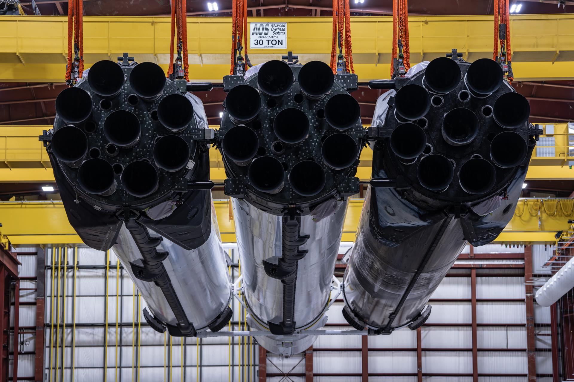 航天图片资料 · A SpaceX Falcon Heavy rocket is lifted by a yellow crane inside an industrial facility. The 27 black rocket engines stick out of the bottom or three white booster housings, some covered with soot.