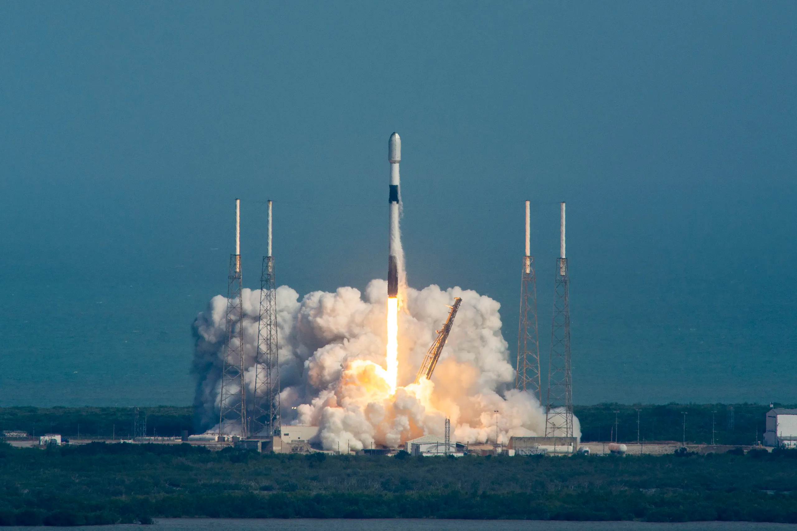 航天图片资料 · A SpaceX Falcon 9 rocket rises from its launch pad at Cape Canaveral Space Force Station, surrounded by a billowing cloud of white exhaust and a bright orange flame at the base. The rocket climbs vertically against a hazy blue-gray sky, flanked by four lightning rod towers. Launch infrastructure, including a yellow crane arm and concrete support structures, is visible at the bottom of the frame.
