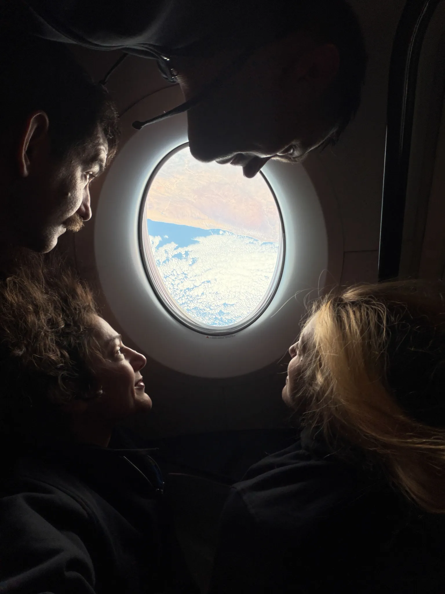 航天图片资料 · NASA’s SpaceX Crew-12 crew members surround an oval-shaped window aboard a SpaceX Dragon, looking out at clouds above blue water and a tan landmass. From the left, rotating clockwise around the image are NASA astronaut Jack Hathaway, Roscosmos cosmonaut Andrey Fedyaev, ESA (European Space Agency) astronaut Sophie Adenot, and NASA astronaut Jessica Meir. Their faces are illuminated by sunlight streaming through the window.