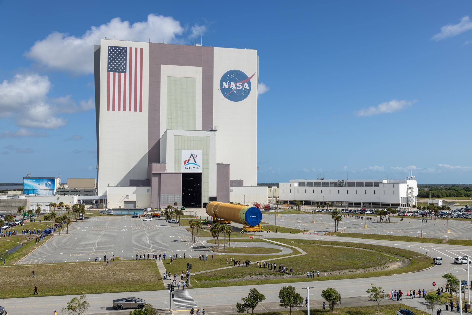 航天图片资料 · The image shows NASA's Kennedy Space Center iconic Vehicle Assembly Building with an American flag painted on the building along with the NASA and Artemis insignias in a blue sky with some clouds on Tuesday, April 28, 2026. People gathered to watch the transport of the Artemis III core stage, an orange yellow that will launch during the Artemis III mission. White buildings, grass, and a parking lot can also be seen in the photo. Photo credit: NASA/Glenn Benson