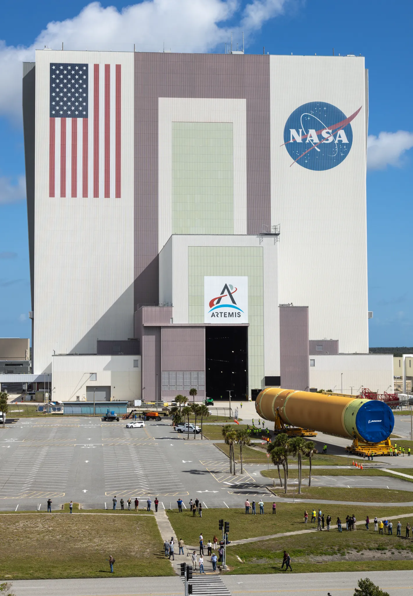 航天图片资料 · The Artemis III rocket core stage, a massive orange cylinder, moves into the tall Vehicle Assembly Building. A line of people watch from afar on a grassy lawn.