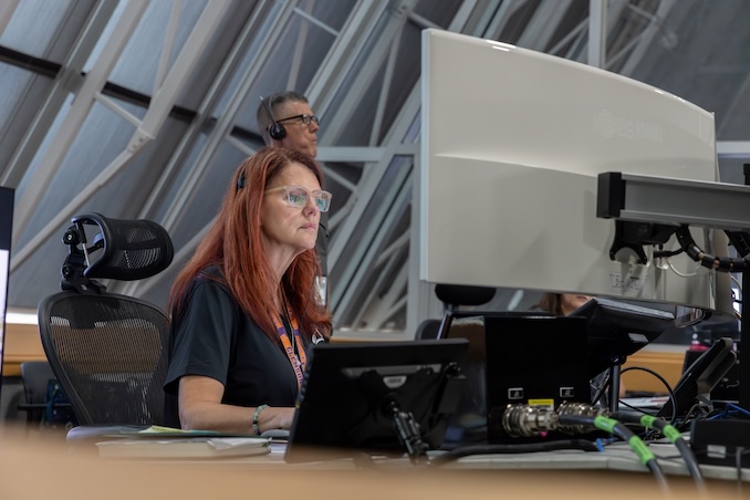 航天图片资料 · Two people wearing headsets work together at computer stations inside a control room setting, with one person seated at a keyboard and the other person standing nearby.