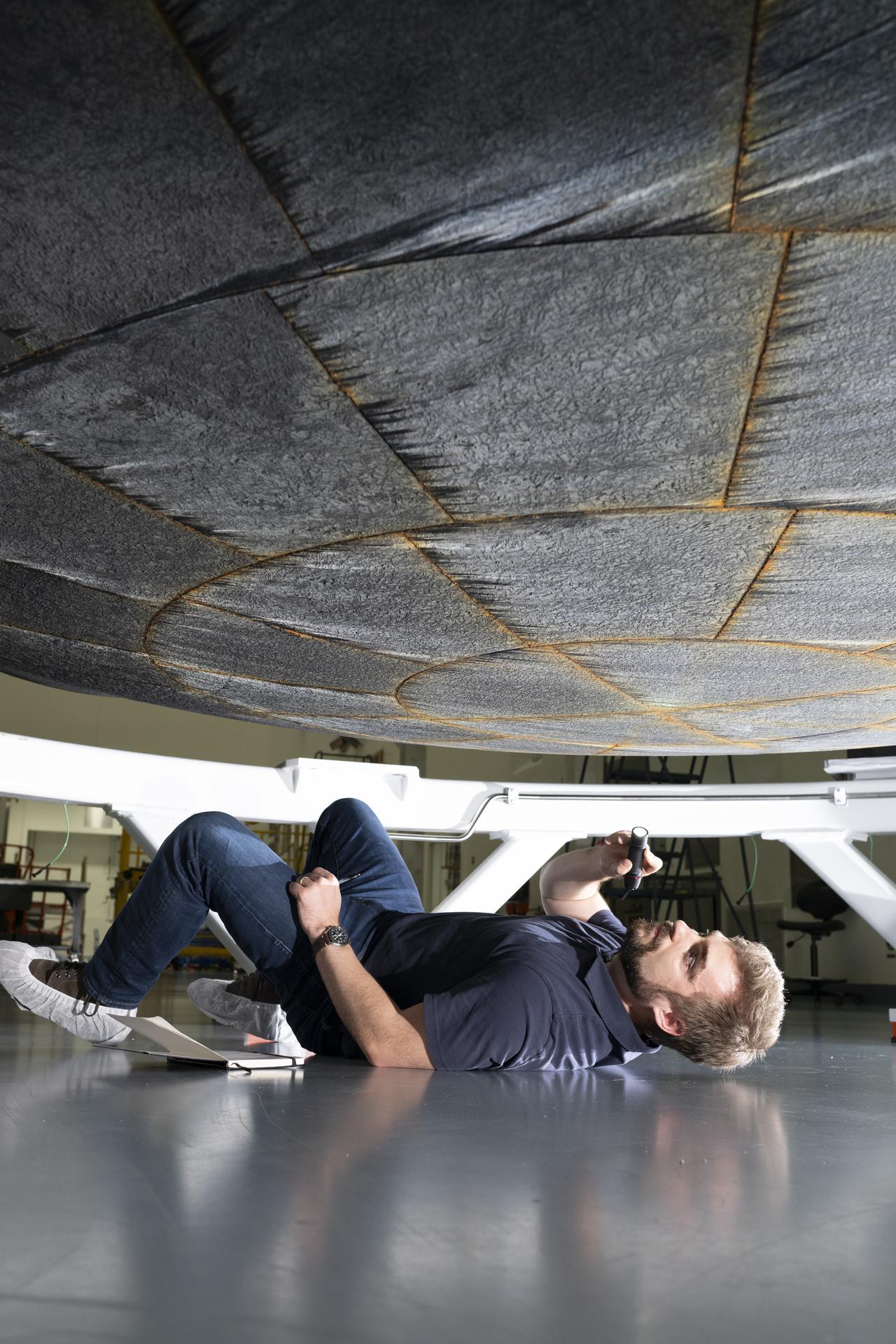 航天图片资料 · A person lies on the floor of a workshop, holding a flashlight to inspect the underside of a large, circular heat-shield-like structure suspended above them. The individual is positioned on their side beneath the hardware, with tools and equipment visible in the background.