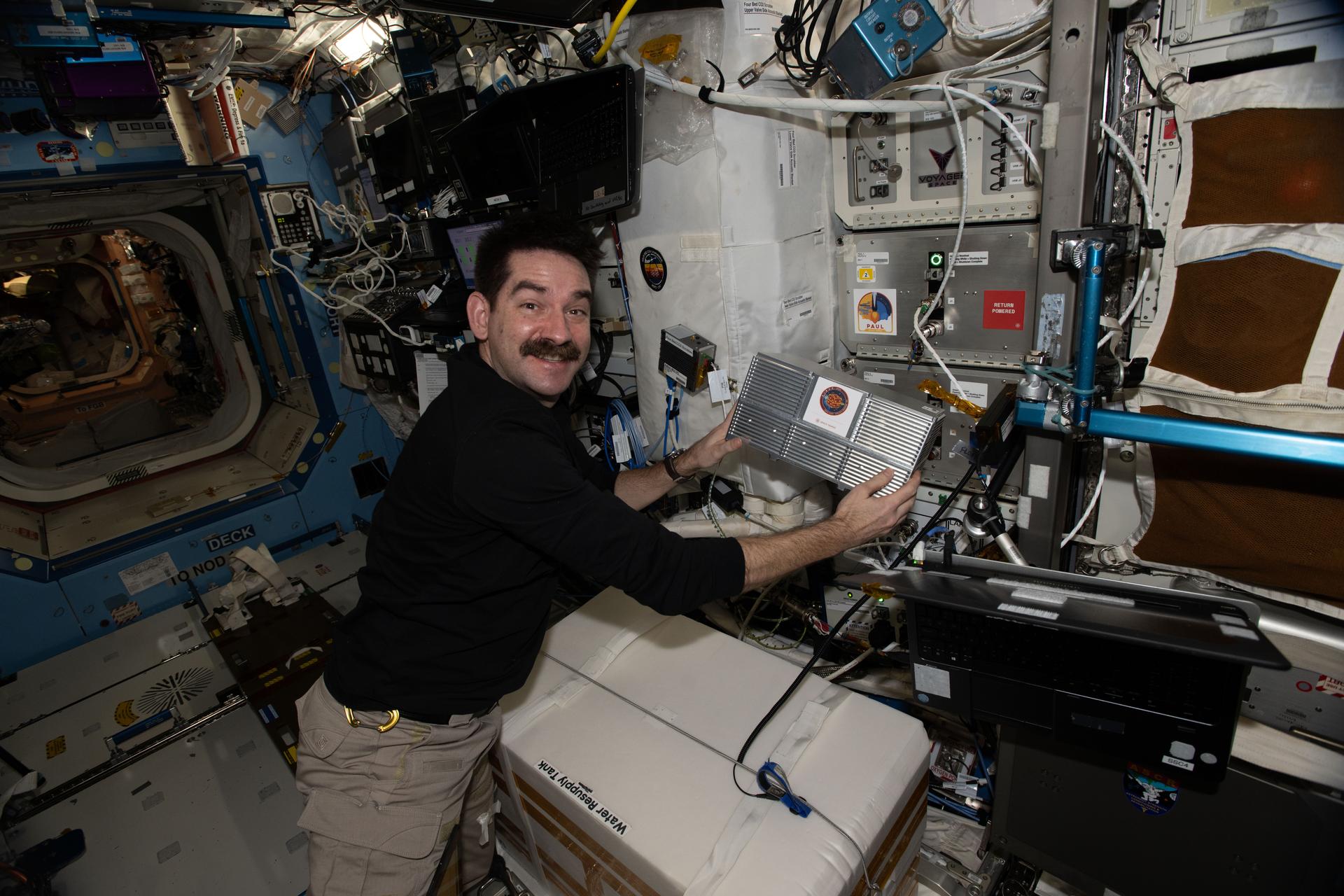 航天图片资料 · NASA astronaut Jack Hathaway smiles while holding a rectangular metallic box in both hands, about the size of a shoe box. There is a large white box in front of him, and an opening to another area of the International Space Station is visible behind him.