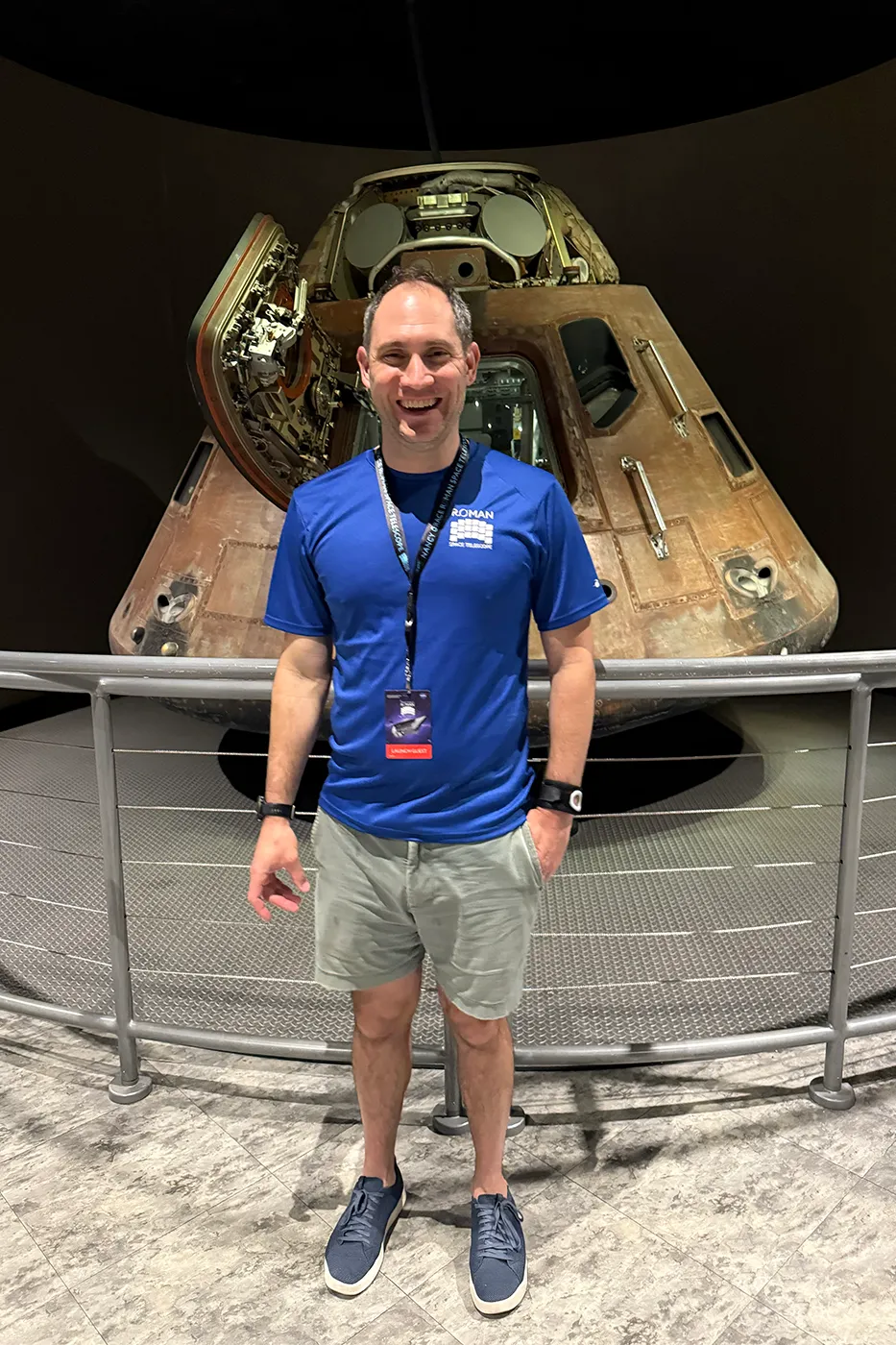 航天图片资料 · A man in a blue T-shirt and shorts smiling in front of a retired space capsule on display.