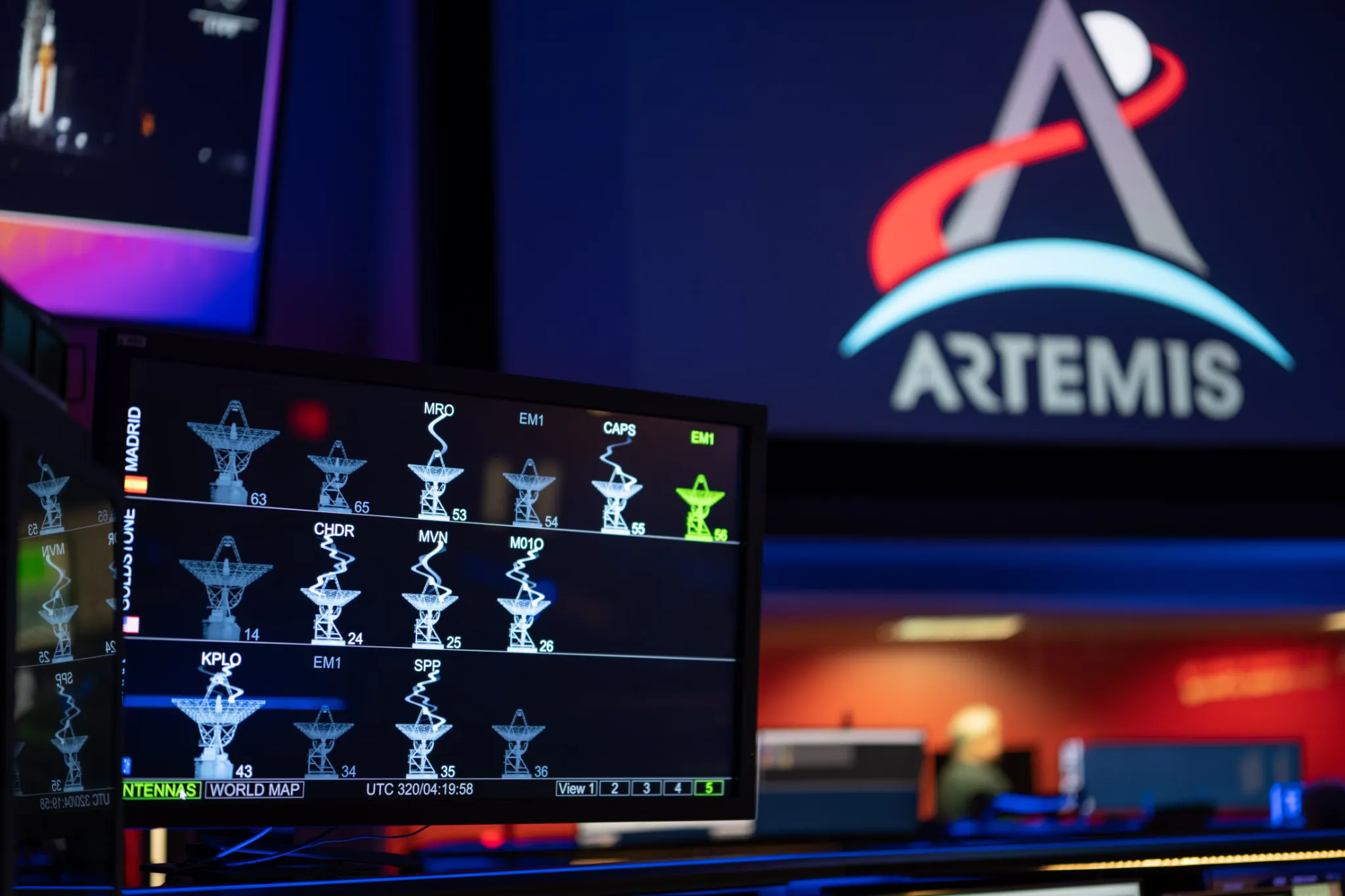 航天图片资料 · The DSN Now tool is displayed on a screen in the foreground—antenna images show real-time data provided by the Deep Space Network ground stations. The Artemis logo is seen in the background on a large screen.