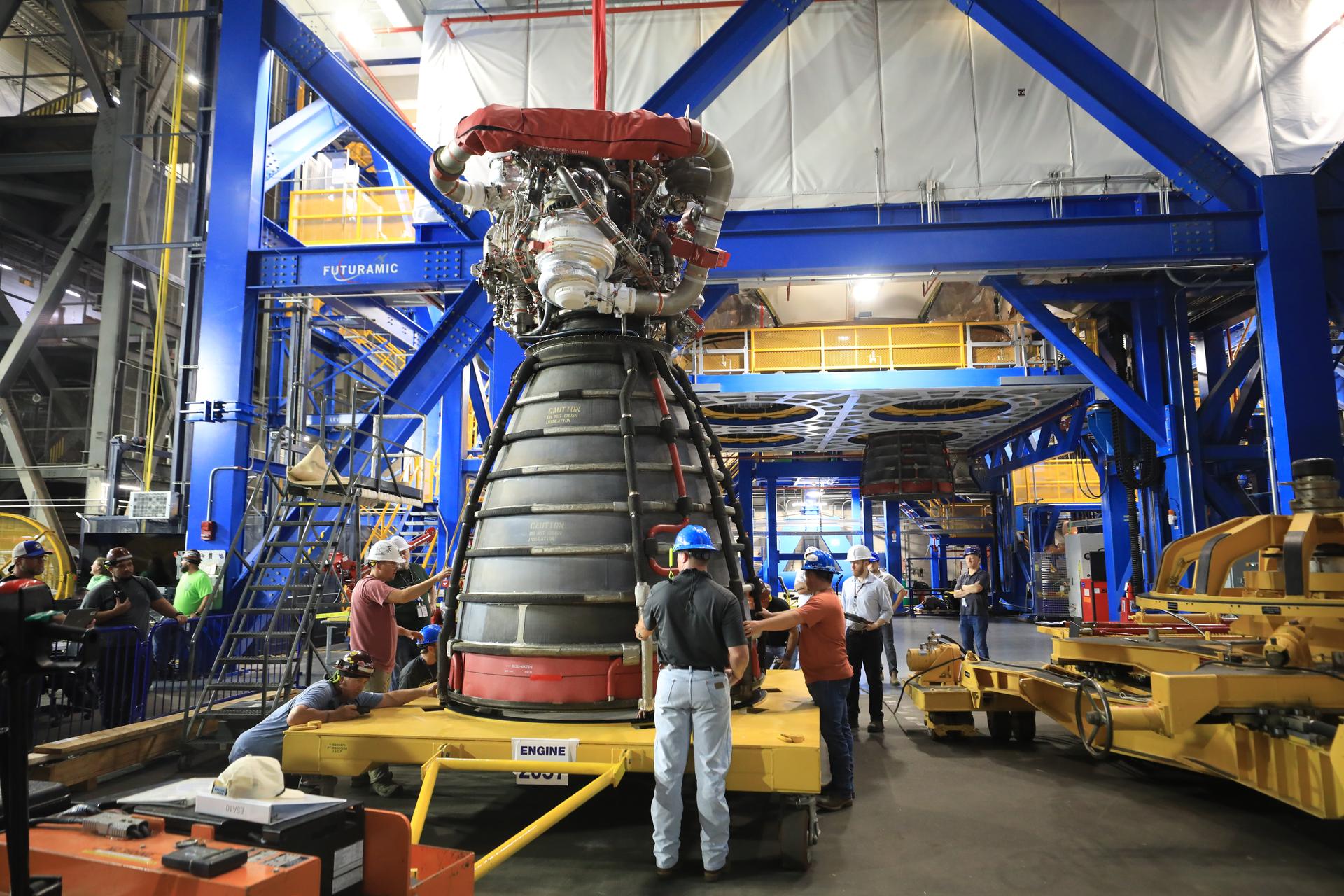 航天图片资料 · A rocket engine sits on a yellow transport platform inside a large industrial facility. Several technicians wearing hard hats and safety gear stand around the engine, inspecting and guiding it. The engine’s bell nozzle faces downward, and complex piping and hardware are visible at the top. Bright blue structural frames and elevated yellow walkways surround the work area, with additional equipment and machinery in the background.