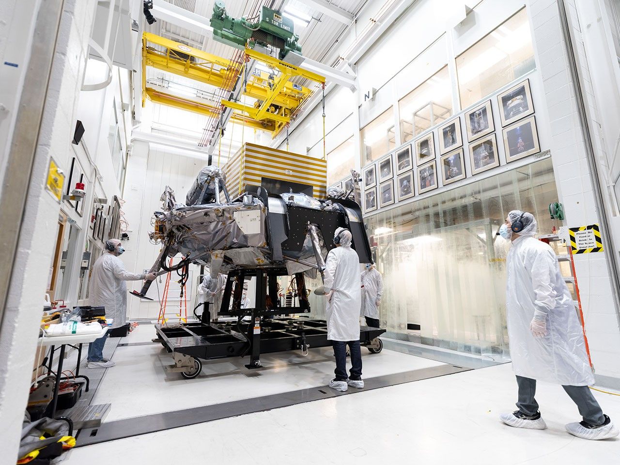 航天图片资料 · Engineers in white clean-room garb monitor a large spacecraft as it is hoisted by a yellow overhead crane in a hallway in a bright, high-tech laboratory. Framed photos line both sides of the hallway.