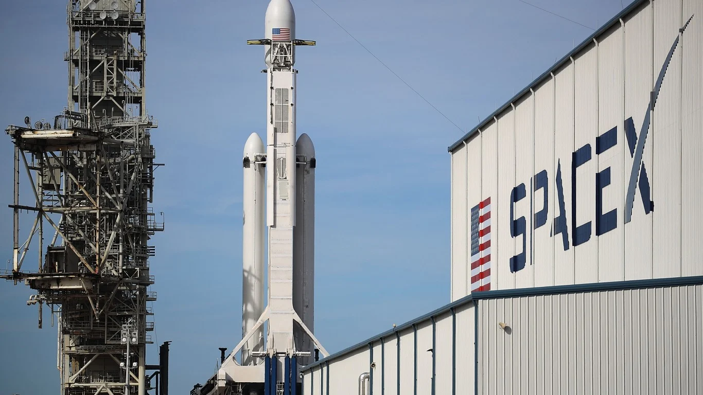 航天图片资料 · A white SpaceX Falcon Heavy rocket, featuring a visible American flag near its nose cone, stands tall on a launchpad. To its left, a complex metal launch tower rises, and to its right, a white corrugated building displays the bold blue 'SPACEX' logo and an American flag design. The background is a bright blue sky.