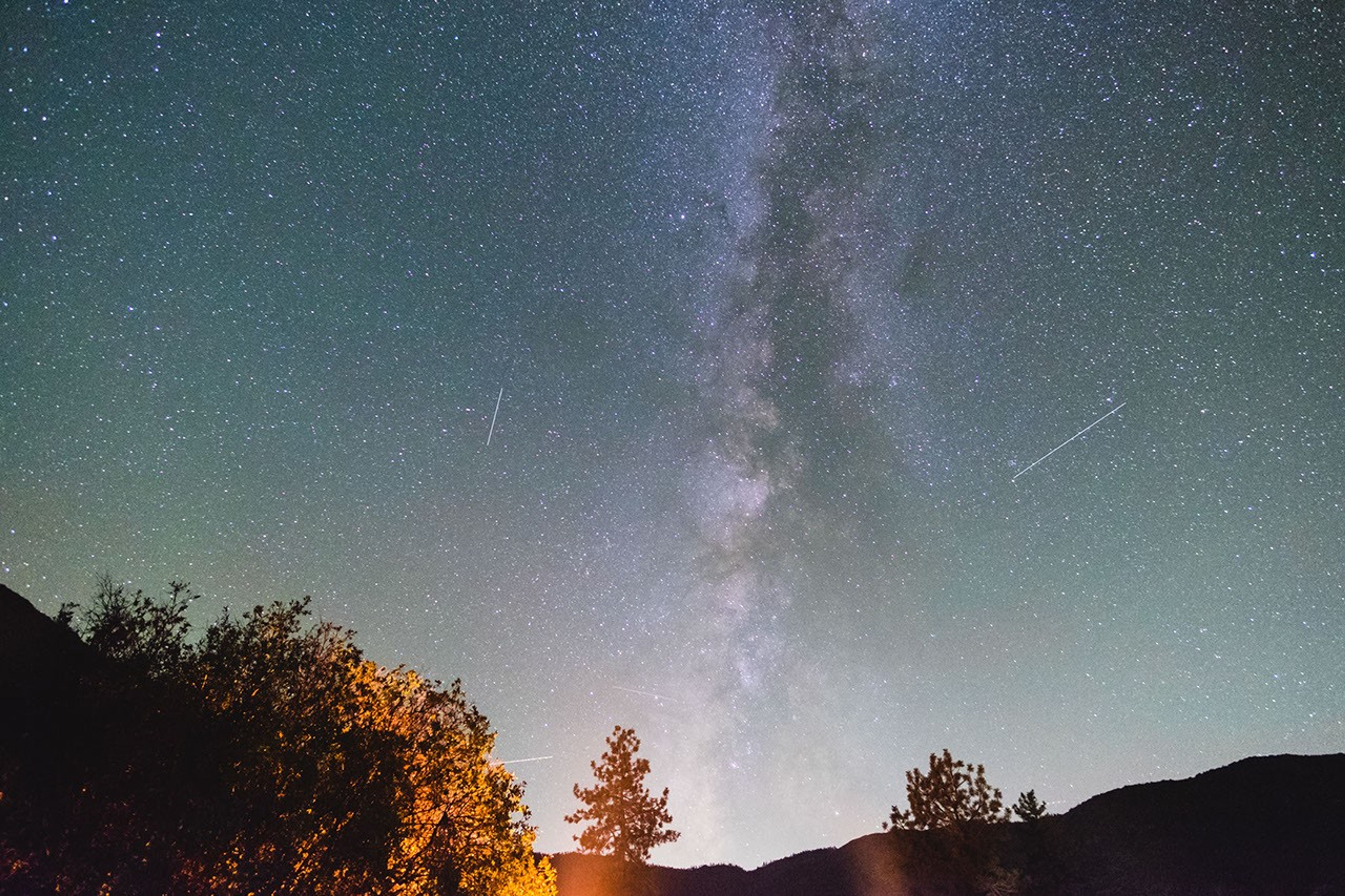 航天图片资料 · A long-exposure photograph of a clear night sky filled with countless stars, featuring the bright, dust-filled band of the Milky Way galaxy stretching vertically through the center. Faint streaks of light from meteors are visible across the sky. Along the bottom, dark silhouettes of trees and distant mountain ridges frame the horizon, with warm light glowing softly behind the foliage on the lower left.