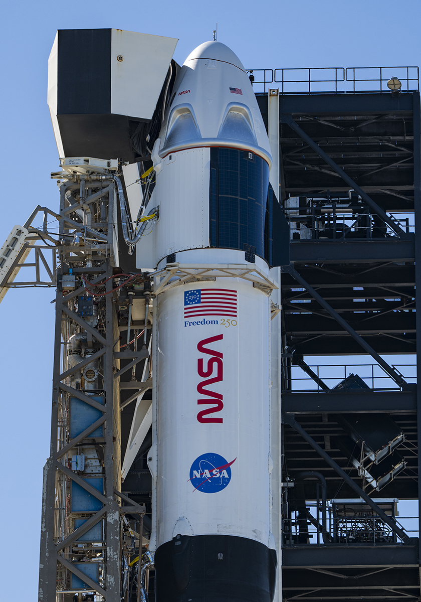 航天图片资料 · A close-up view of a SpaceX Crew Dragon spacecraft mounted atop a Falcon 9 rocket on the launch pad. The capsule and trunk are connected to ground support hardware, with NASA and mission insignia visible on the rocket’s exterior against a bright blue sky.