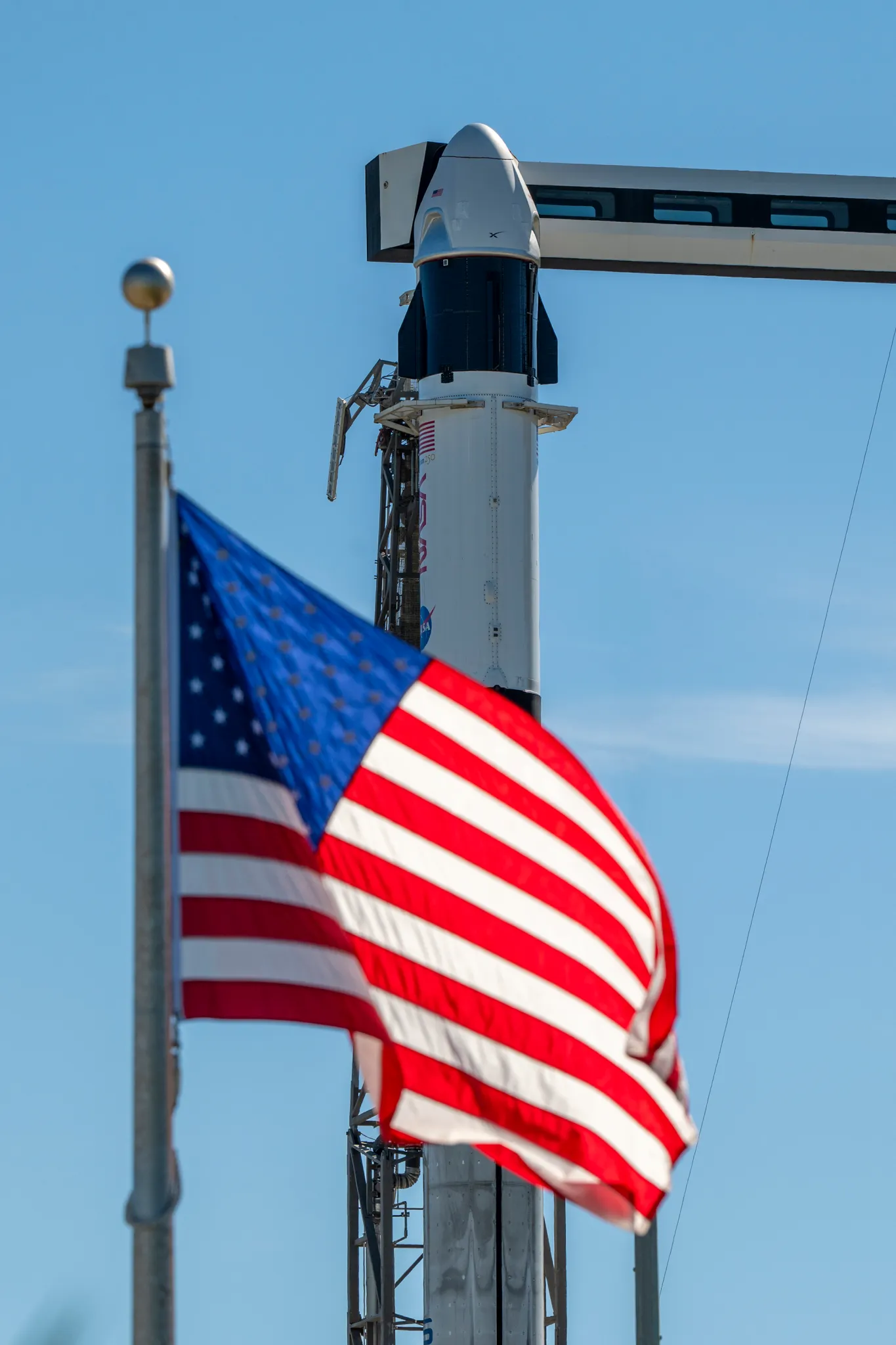 航天图片资料 · A close-up view of a SpaceX Crew Dragon spacecraft mounted atop a Falcon 9 rocket on the launch pad. The capsule and trunk are connected to ground support hardware, with NASA and mission insignia visible on the rocket’s exterior against a bright blue sky. in the foreground in the American flag.