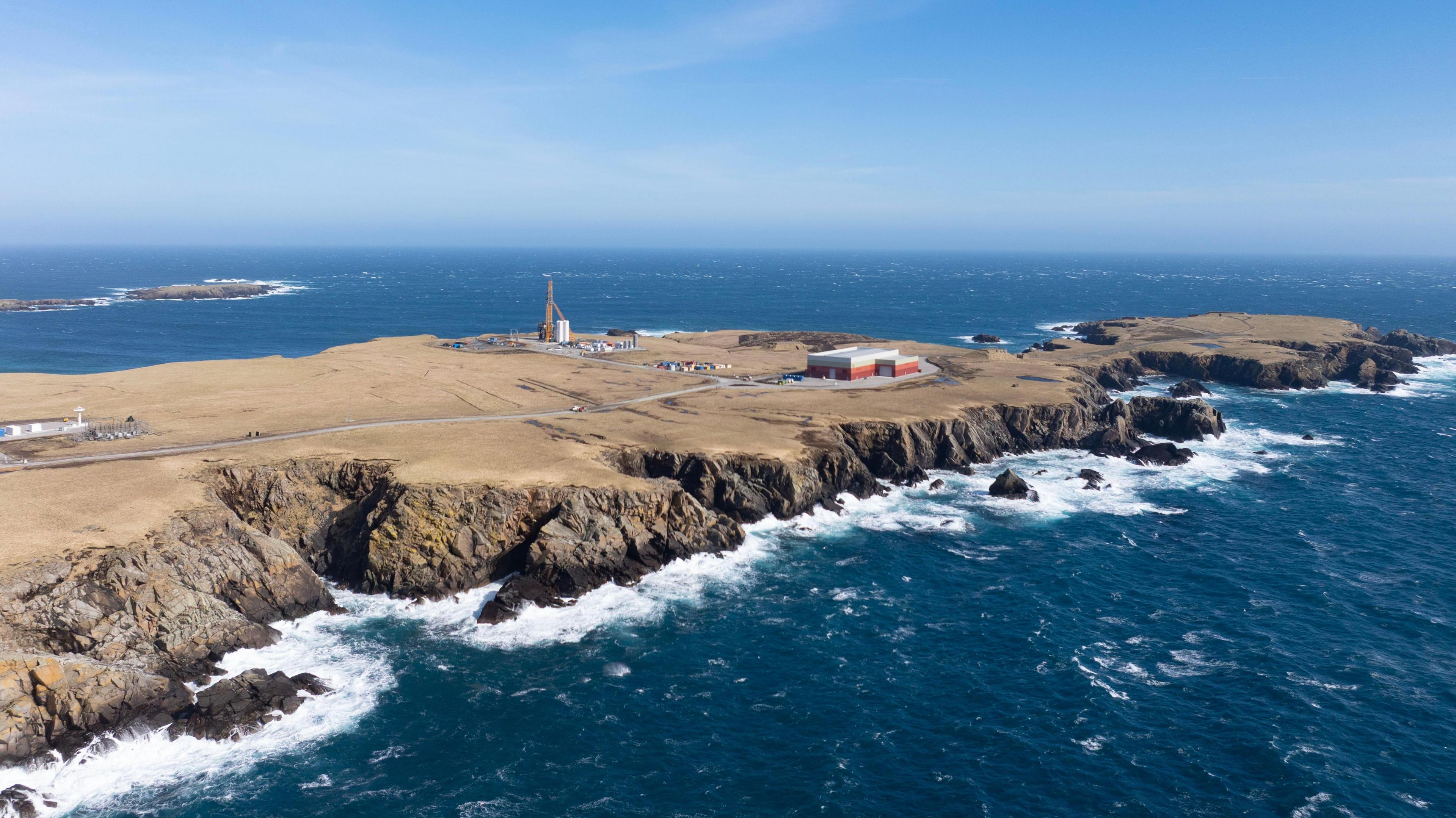 航天图片资料 · An aerial view of a long flat peninsula surrounded by dark blue sea. The coastline is rocky and the sky is blue. The island is brown and sparsely populated by several industrial buildings and a tall launch pad.