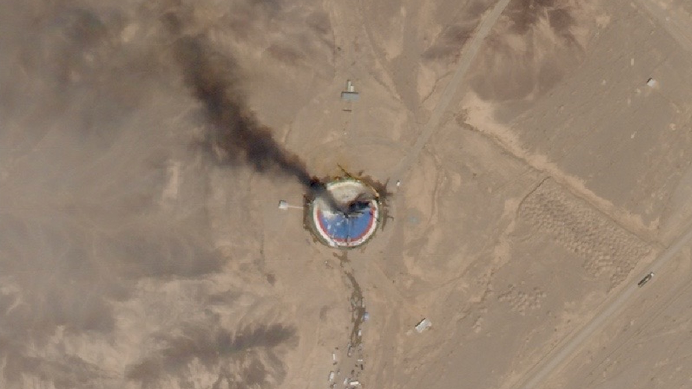 航天图片资料 · satellite photo of a dark plume of smoke ascending from a blue and red circular launch pad in a desert landscape
