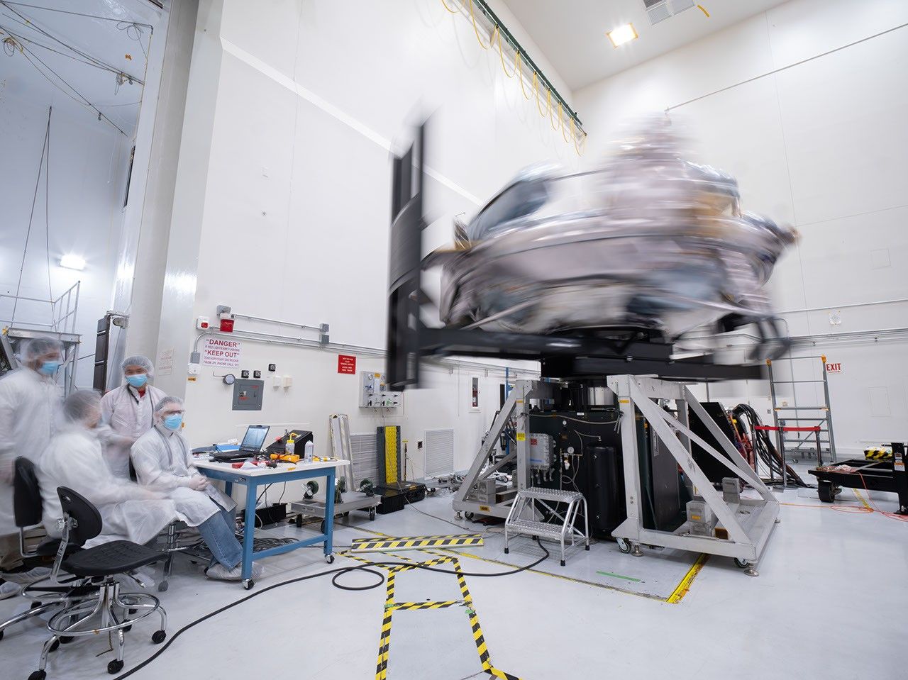 航天图片资料 · Engineers in white clean-room frocks monitor a silvery spacecraft spinning so rapidly it’s blurred on a test rig inside a bright laboratory.