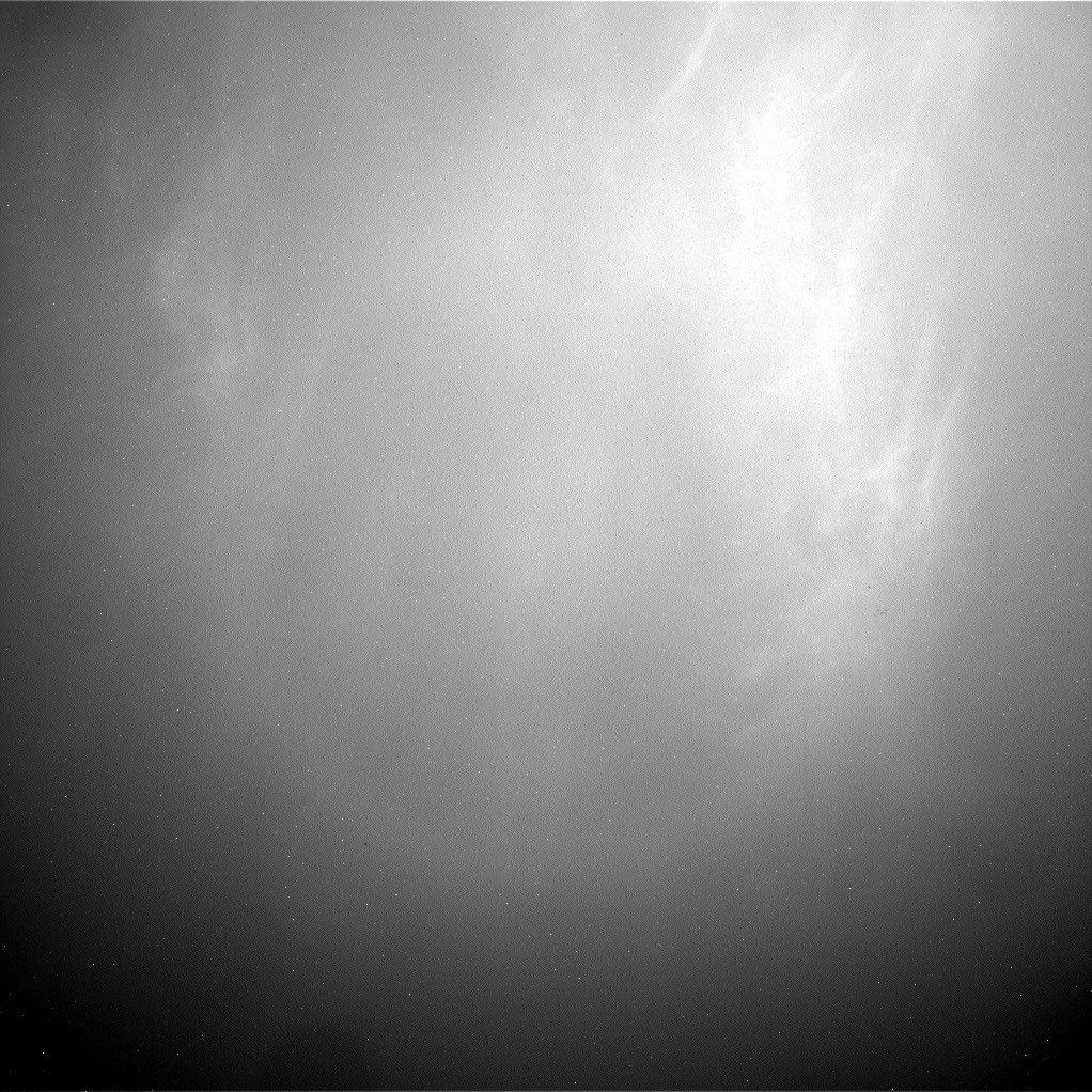 航天图片资料 · A grainy, black-and-white image of the sky above Mars, captured by the Curiosity rover. The upper right portion of the image is brightly illuminated, highlighting delicate, wispy, cirrus-like cloud structures. The brightness diffuses outward, transitioning into a dark, textured gradient toward the bottom and left edges of the frame.