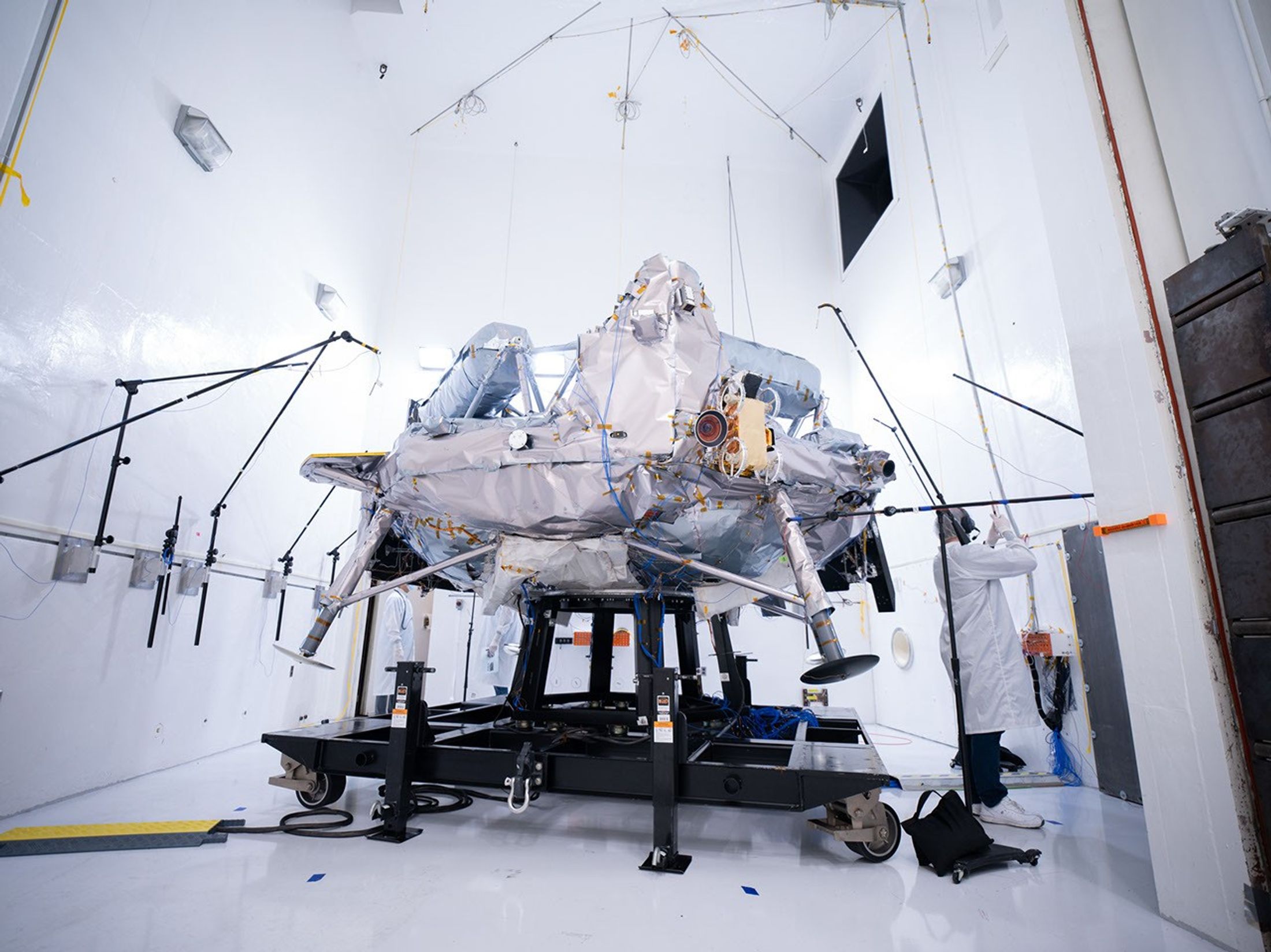 航天图片资料 · A silvery spacecraft sits on a test frame surrounded by microphones and testing equipment inside a bright white acoustic test chamber, while an engineer in a lab coat works nearby.