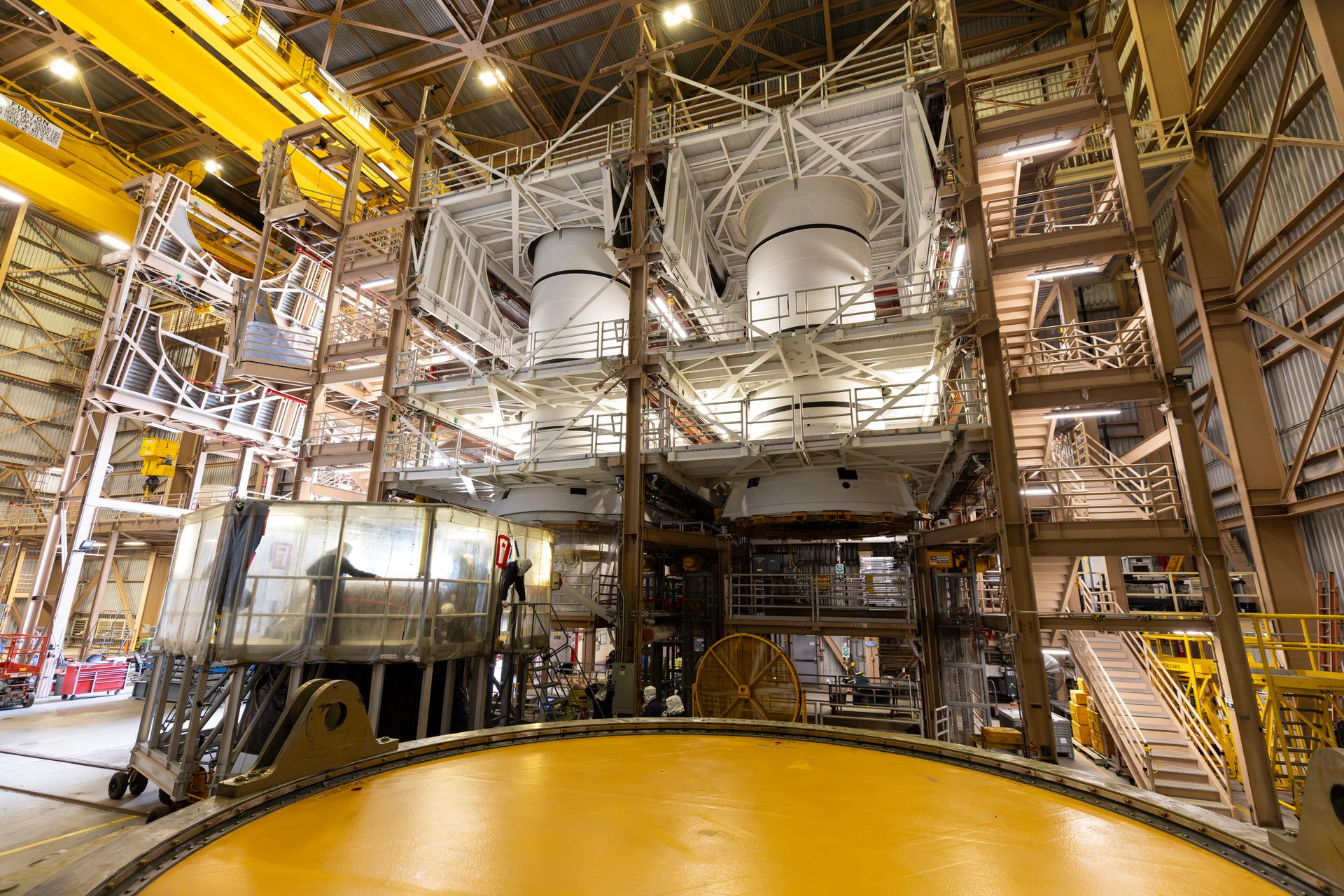 航天图片资料 · Inside a large industrial facility, two massive rocket engines or tank structures are suspended high within a metal scaffolding system. The surrounding area is filled with stairways, platforms, pipes, and support beams. In the foreground, a circular yellow platform dominates the lower part of the scene. Overhead lights illuminate the spacious interior, highlighting the complex machinery and structural framework.
