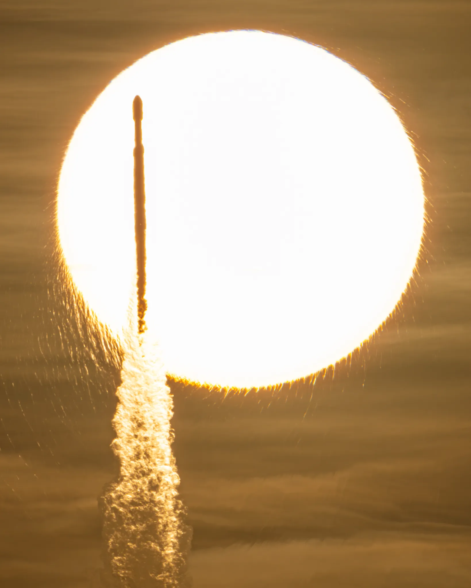 航天图片资料 · In a sepia-toned photo, the Sun appears as a bright, enormous orb that appears to have frayed edges. Against this backdrop, a tall and slender rocket is seen in silhouette atop a cloudy plume.