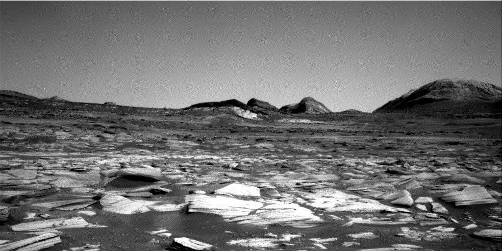 航天图片资料 · A grayscale image of the Martian surface taken by the Curiosity rover. The immediate foreground features a dense field of jagged, light-colored, thinly-layered rock fragments resting on a bed of darker sand or dust. The barren terrain gently slopes upward toward the horizon, where a cluster of dark, rugged hills and a larger, rounded mountain peak on the far right stand out against a smooth, featureless sky.