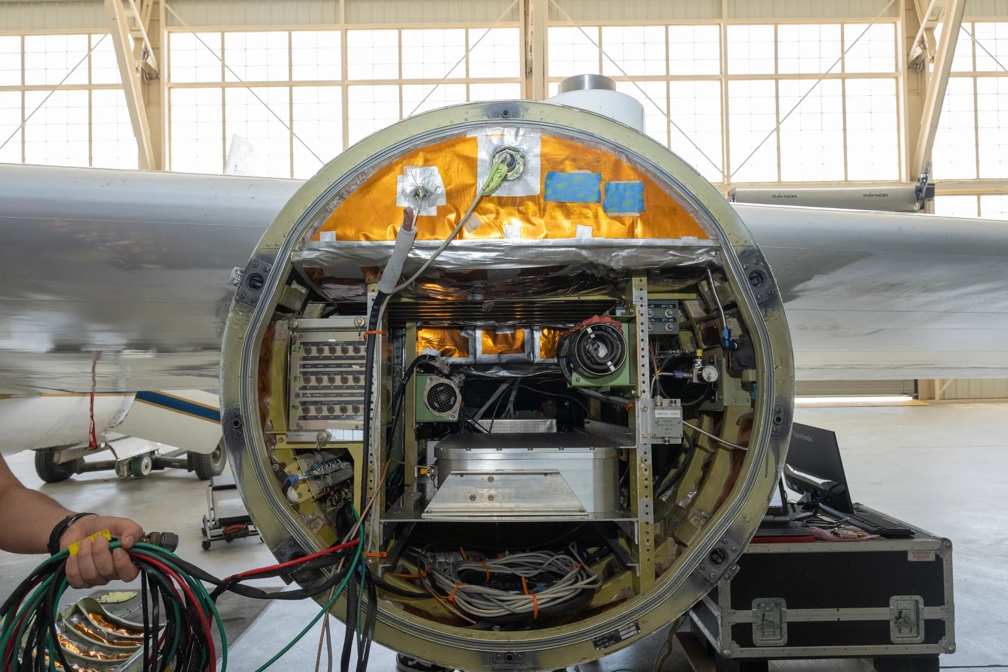 航天图片资料 · Front view of an open instrument pod mounted beneath a NASA ER‑2 aircraft, showing cables, sensors, and electronic equipment arranged inside a circular housing. A person’s hand holding a bundle of cables is visible on the left side, with the aircraft hangar visible in the background.