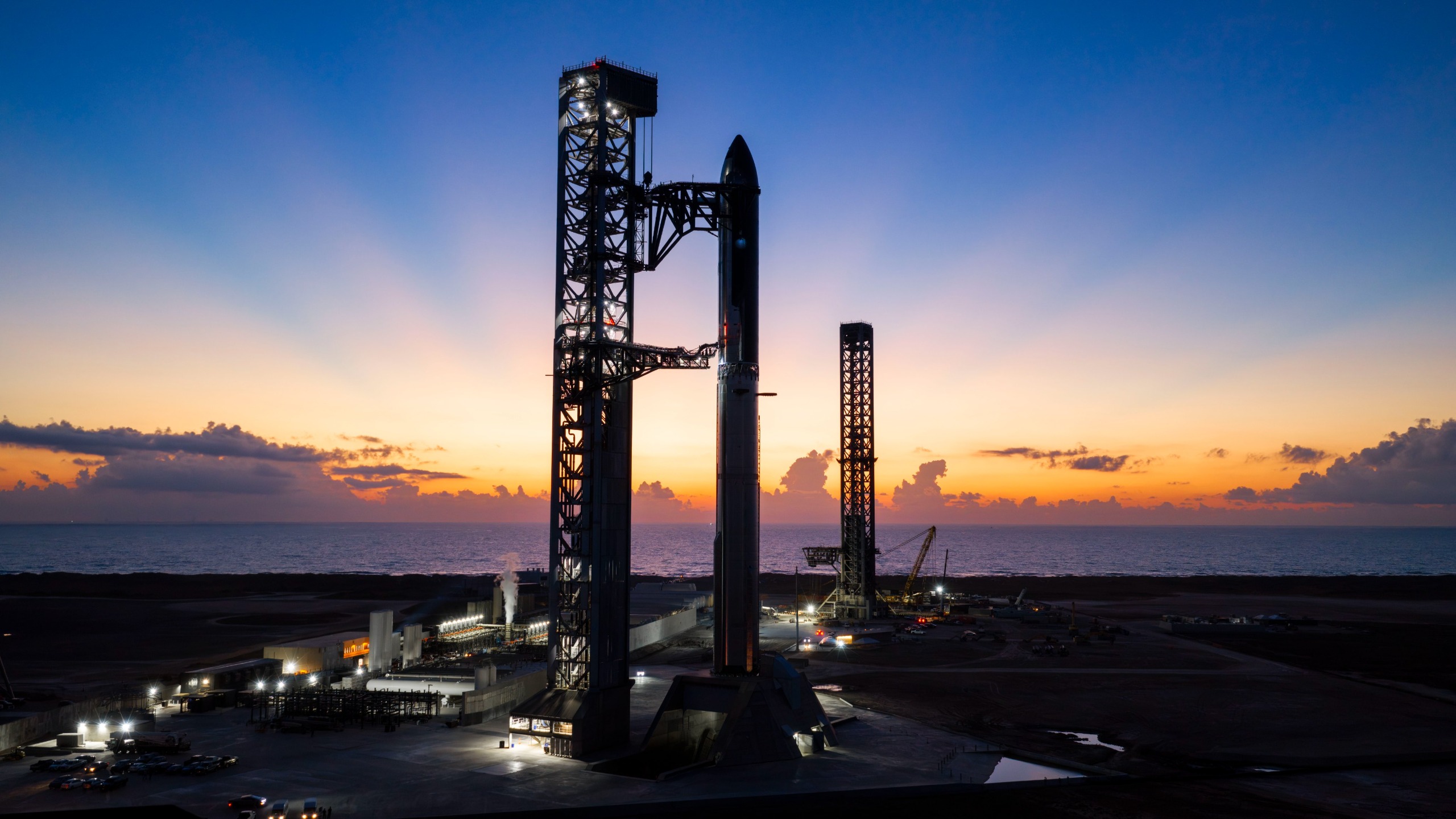 航天图片资料 · A towering SpaceX Starship rocket stands at Starbase's Pad 2, silhouetted against a dark blue sky and yellow rays from a rising sun.