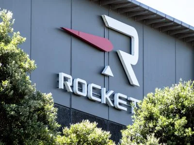 航天图片资料 · A close-up shot of the Rocket Lab company logo and name on a dark grey building facade. The logo features a large white stylized 'R' symbol with a red arrow-like shape pointing left next to it, and a small white triangle beneath the 'R'. Below the logo, the word 'ROCKET' is spelled out in bold, white, 3D letters. Green bushes with sunlit leaves partially cover the lower part of the sign and building.