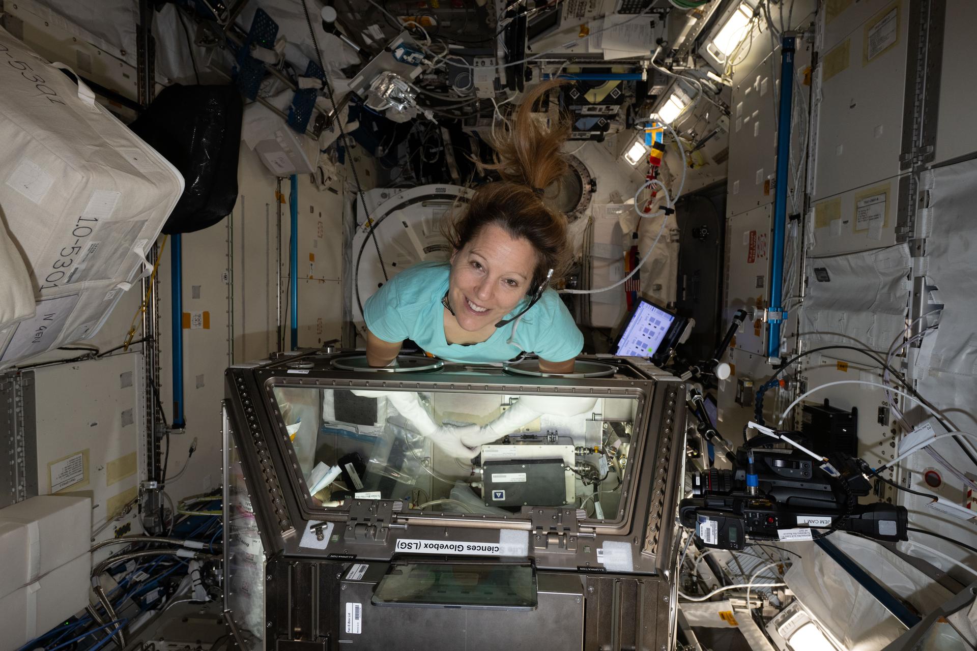 航天图片资料 · ESA (European Space Agency) astronaut Sophie Adenot smiles at the camera as she reaches inside the large, rectangular Life Sciences Glovebox aboard the International Space Station. Her ponytail floats above her head as she leans into the open compartment, surrounded by reflective silver panels, cables, tubing, and equipment.