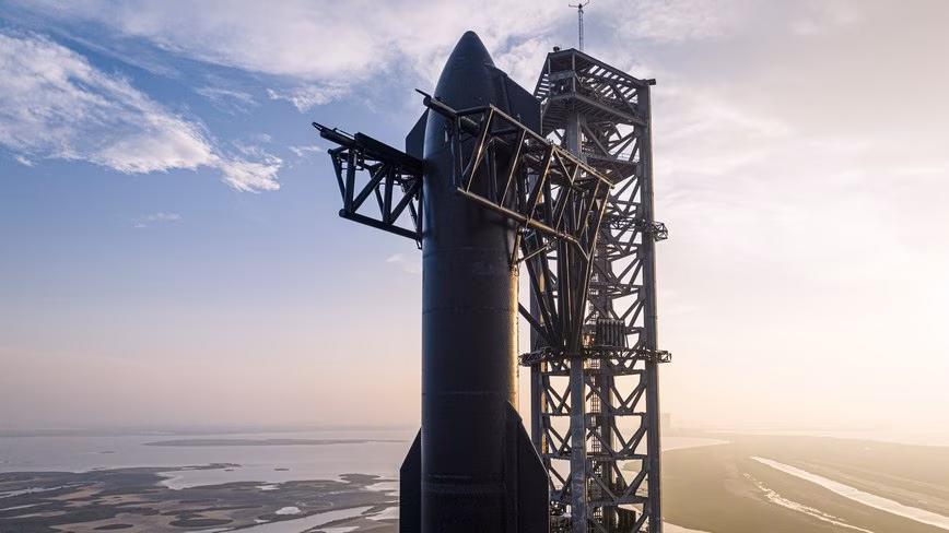 航天图片资料 · Starship on the launch tower at Starbase SpaceX's Starship rocket standing beside its launch tower at Starbase, Texas, with coastal wetlands below at sunrise