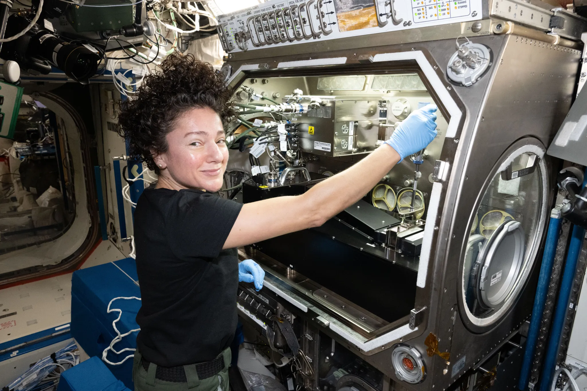 航天图片资料 · NASA astronaut Jessica Meir smiles at the camera in front of the rectangular Microgravity Science Glovebox aboard the International Space Station. Wearing blue gloves, she reaches into the open compartment, which contains experiment hardware surrounded by metallic panels, cables, and tubing.