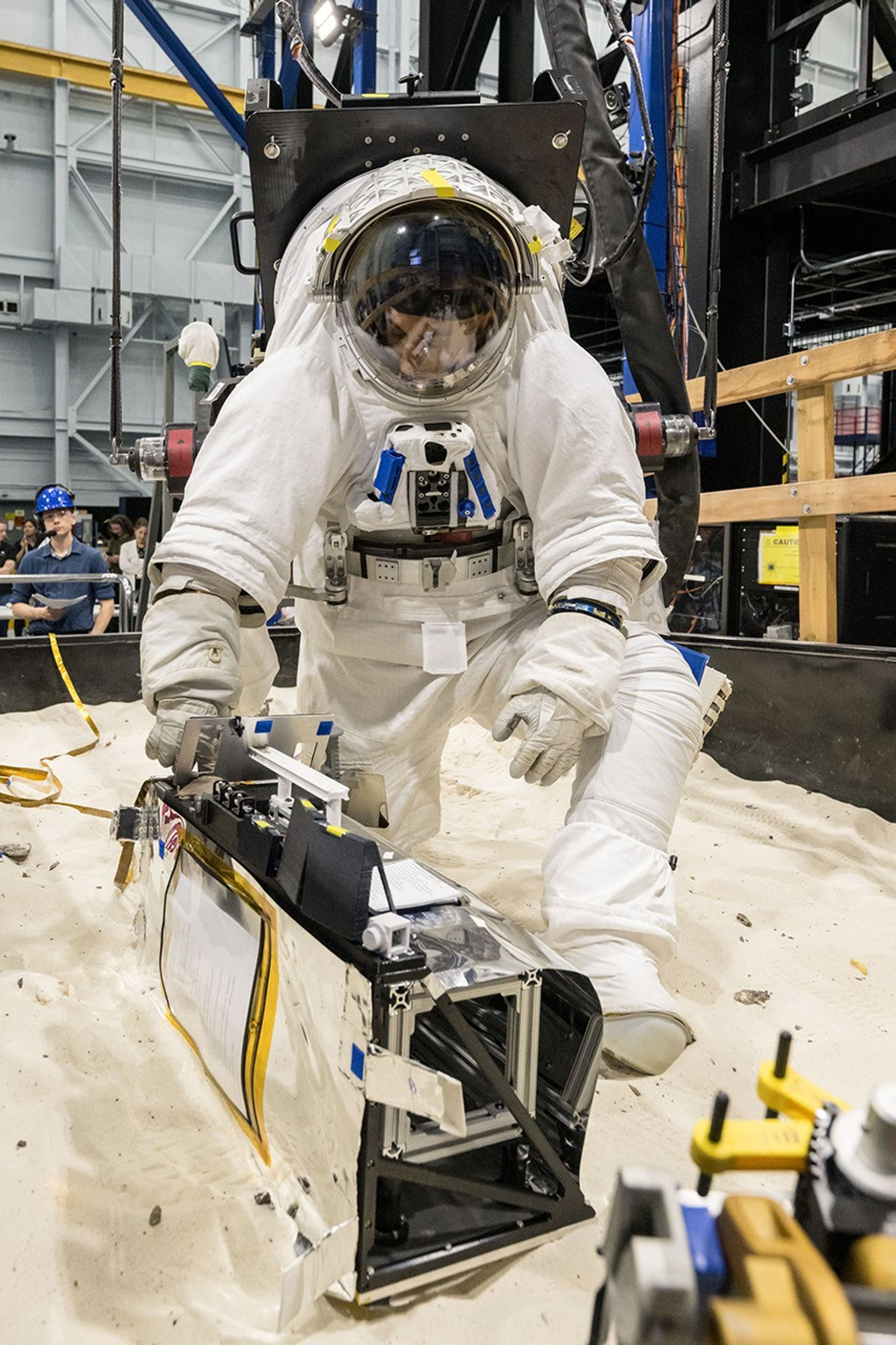 航天图片资料 · An astronaut in a white spacesuit kneels in simulated lunar soil while working with scientific equipment in a large testing facility. Staff members and support structures are visible in the background.