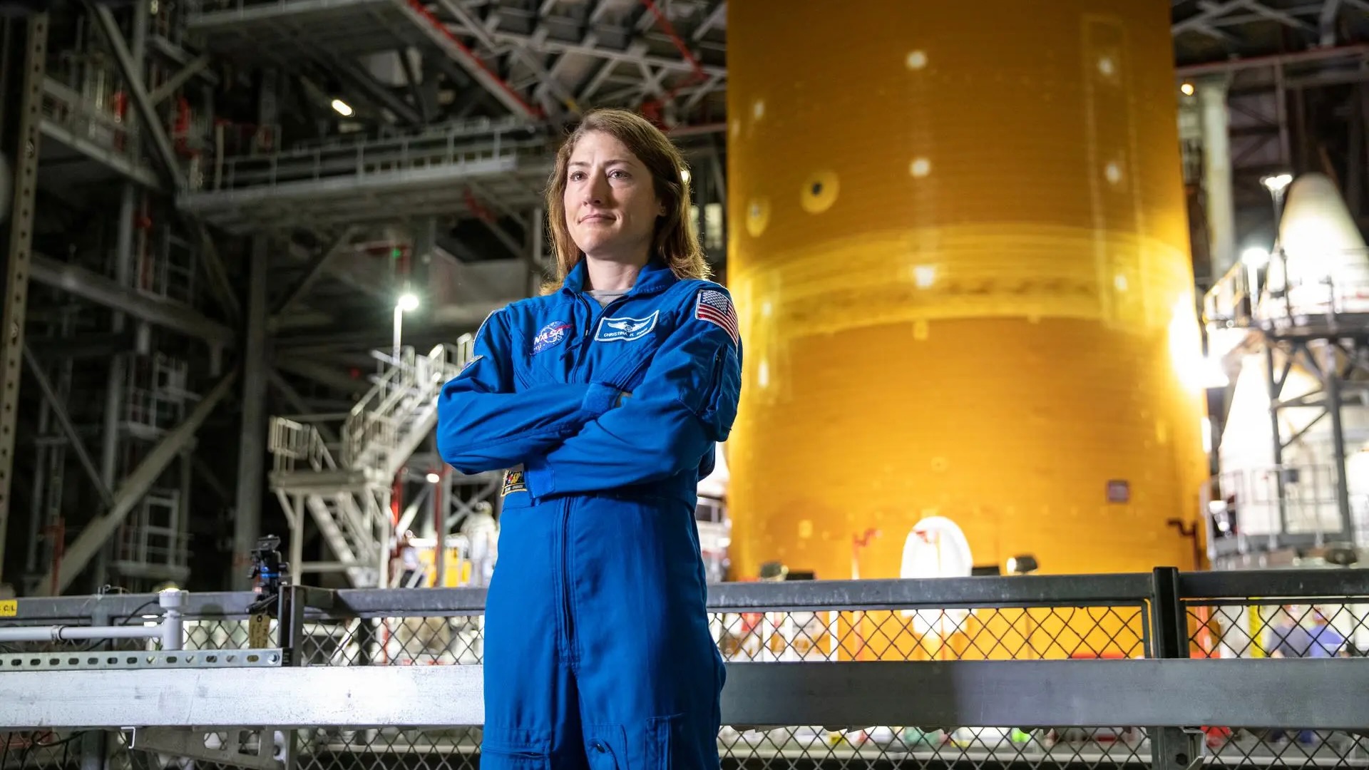 航天图片资料 · a woman in a blue flight suit stands in front of a large orange rocket inside a large building