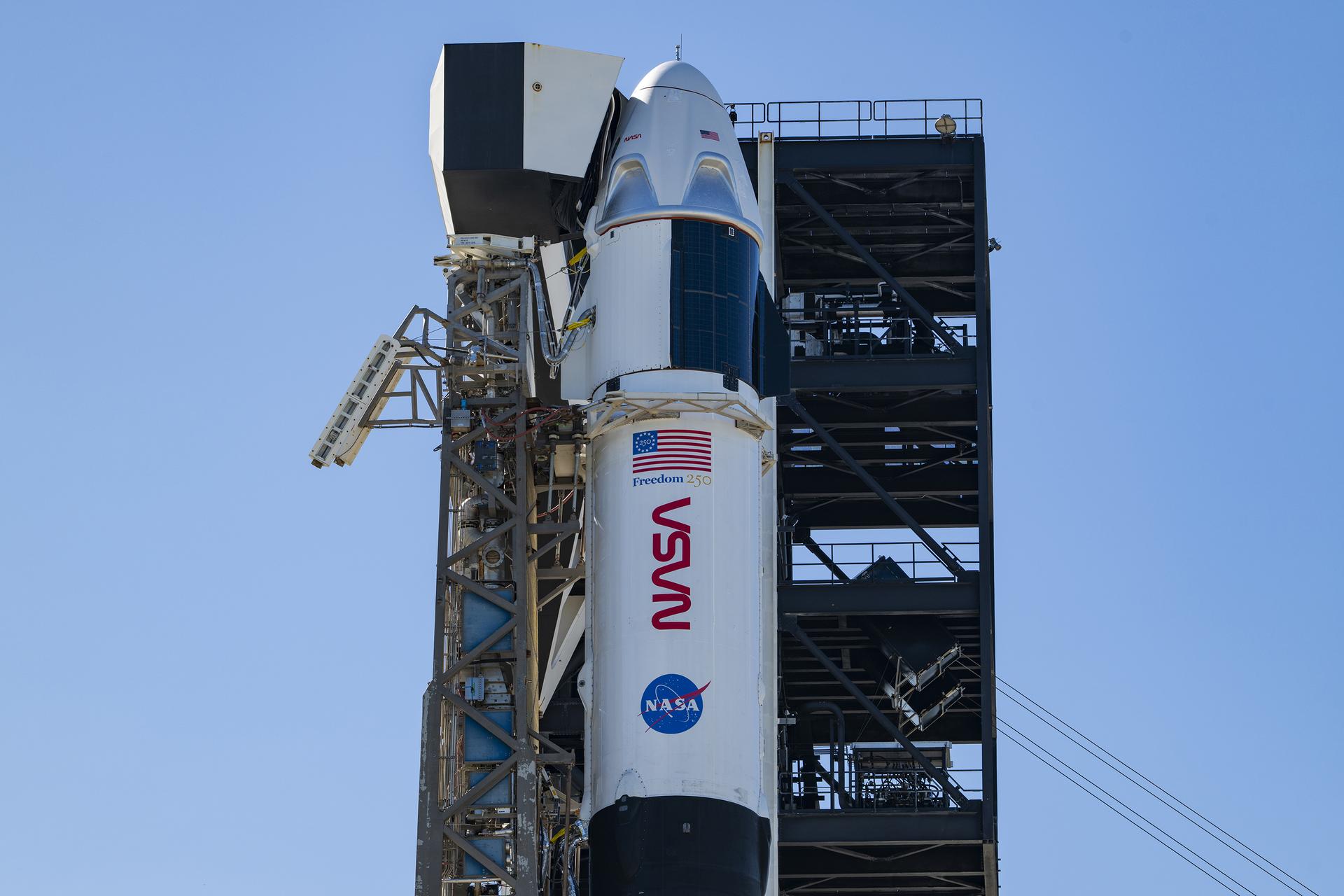 航天图片资料 · A close-up view of a SpaceX Crew Dragon spacecraft mounted atop a Falcon 9 rocket on the launch pad. The capsule and trunk are connected to ground support hardware, with NASA and mission insignia visible on the rocket’s exterior against a bright blue sky.