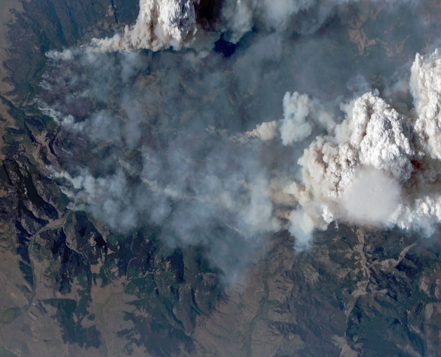 航天图片资料 · Natural-color aerial image of a wildfire in mountainous terrain. Thick gray and brown smoke spreads across much of the landscape, while a towering white pyrocumulus cloud rises at right. A smooth, thin pileus cloud hovers just below the main convective cloud.