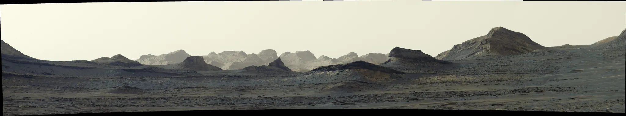 航天图片资料 · A broad, panoramic view of a stark, grayish-tan Martian landscape features jagged rock formations and rolling mountain ridges beneath a hazy sky. Features in the front are darker while distant rock formations are washed in pale light.