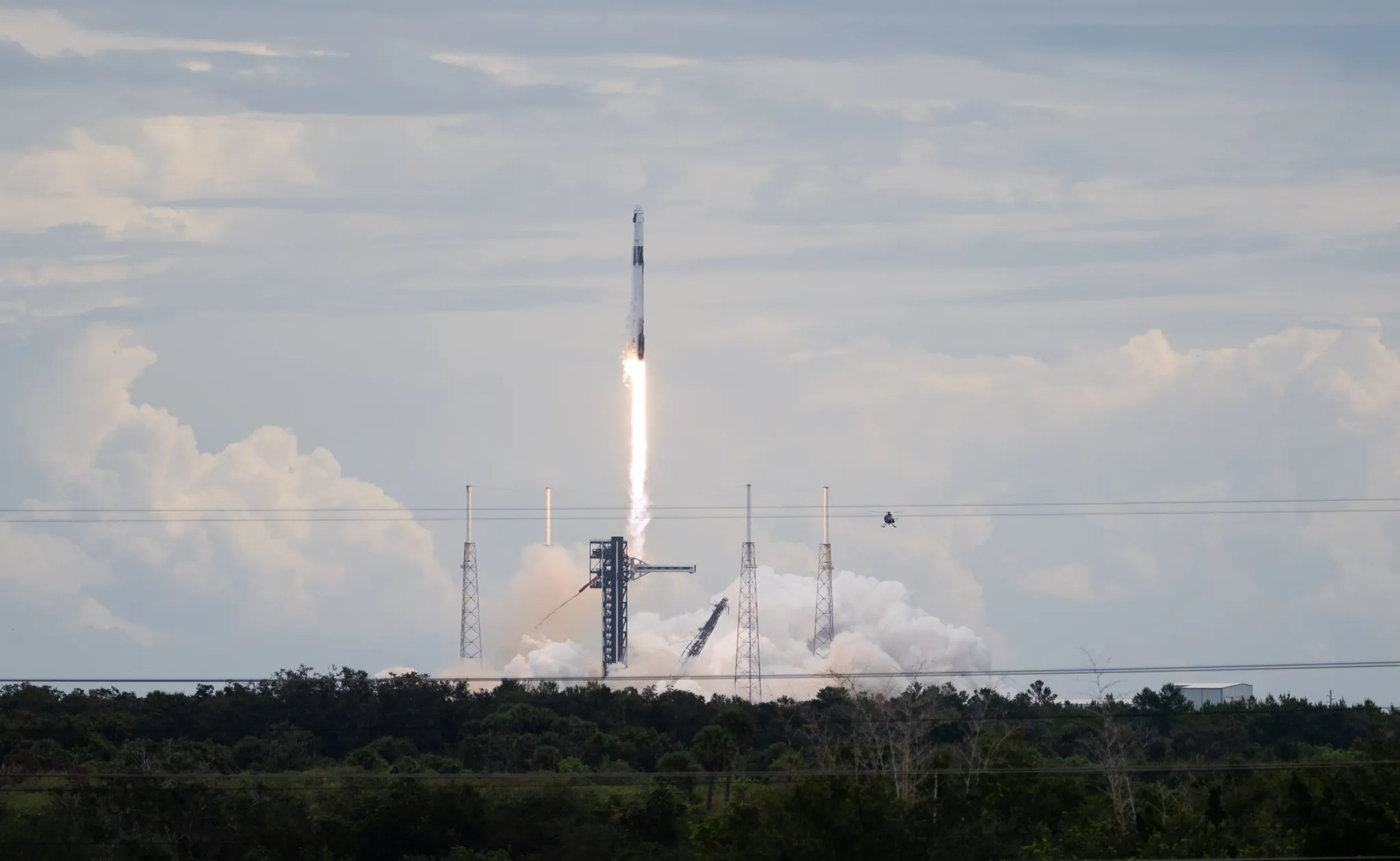 航天图片资料 · A SpaceX Falcon 9 rocket carrying the company's Dragon spacecraft launches. There is a bright column of fire under the rocket as it moves upward. Clouds of vapor spread out below the rocket, almost blending into the similarly shaped clouds in the sky.