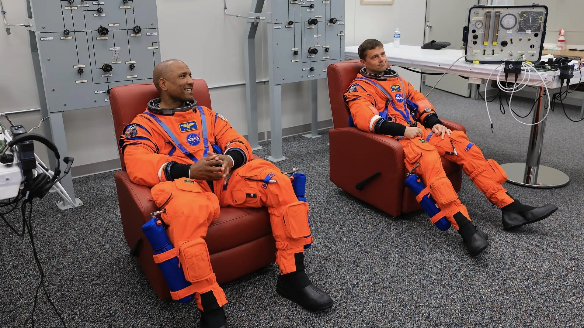 航天图片资料 · two male astronauts in orange flight suits sit in reddish recliners inside a white-walled room