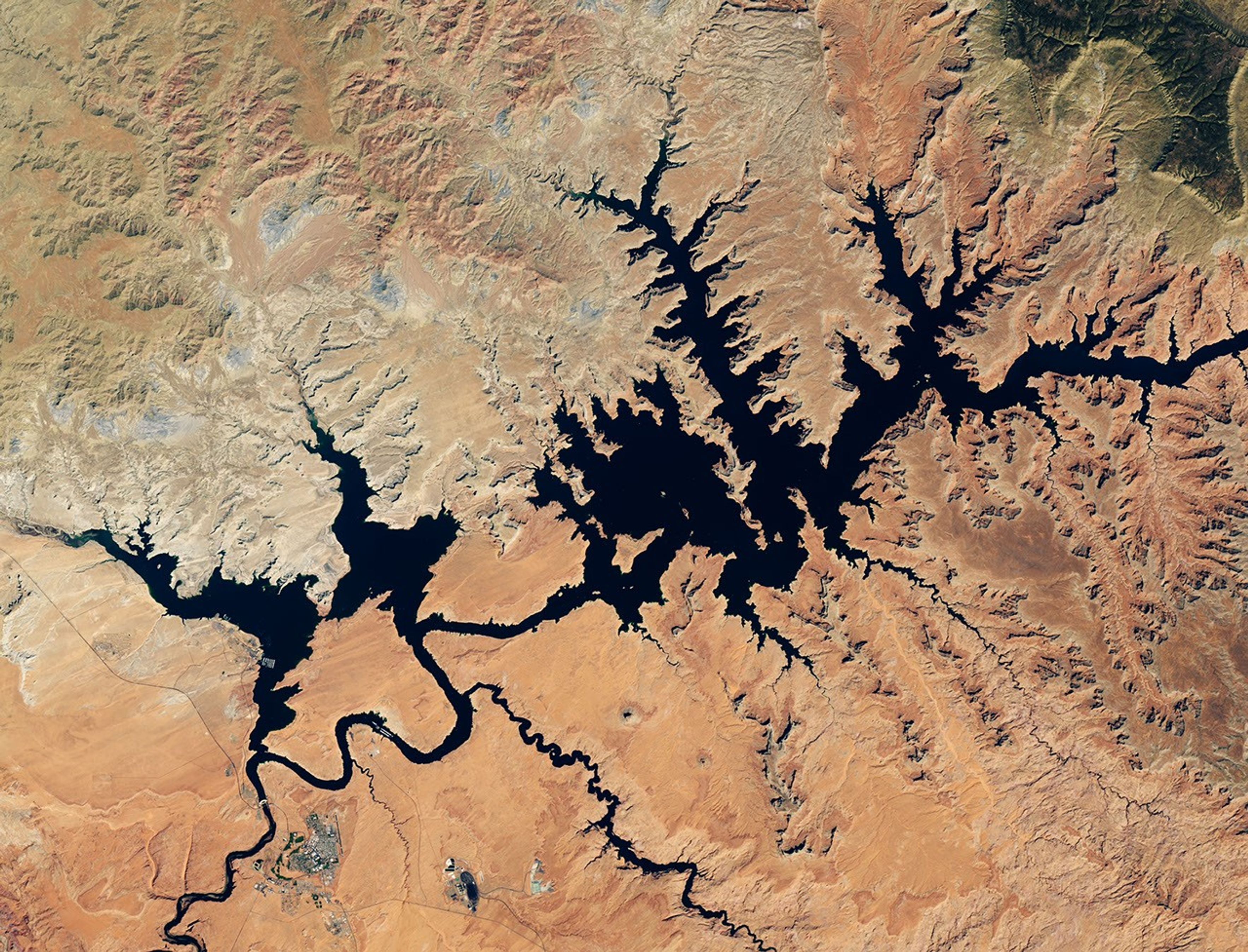 航天图片资料 · Dark blue water in Lake Powell fills several bays and branching canyons amid a light orange desert landscape. The Glen Canyon Dam and the town of Page, Arizona, are visible in the lower left.