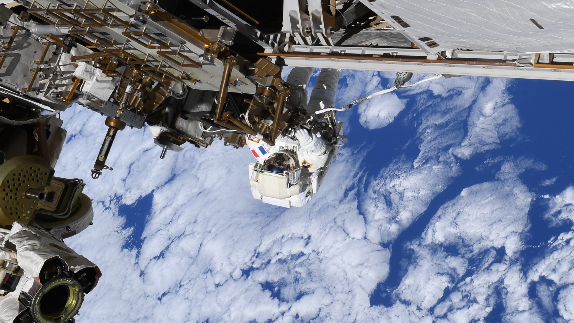 航天图片资料 · a person in a bulky white spacesuit clings to metal bars attached to a large structure, while white clouds in a blue sky can be seen far below
