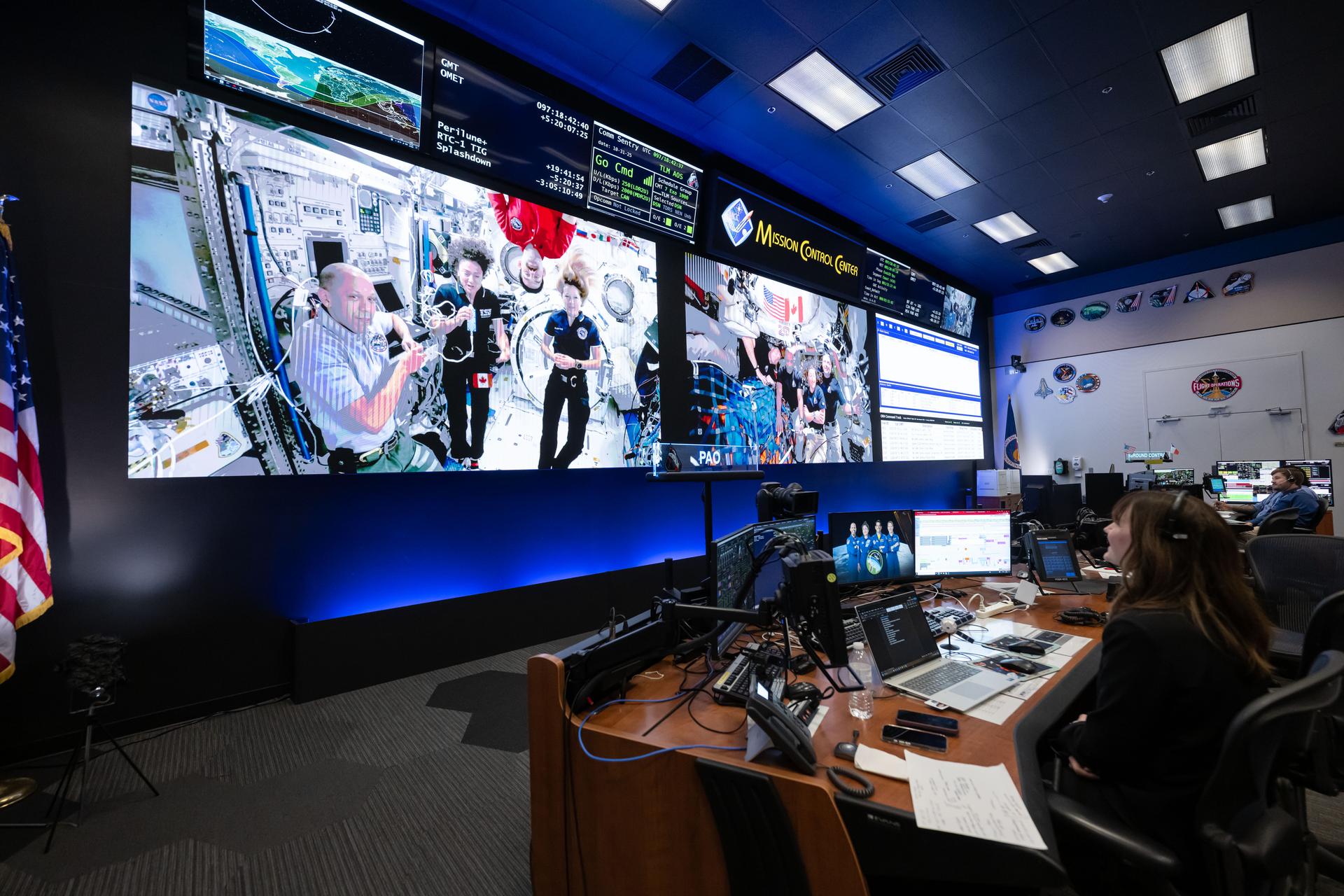 航天图片资料 · Two screens at the front of Mission Control show three astronauts floating at the International Space Station (left) and four astronauts floating in the Orion spacecraft (right). Flight controllers watch from behind computer screens at their desks.