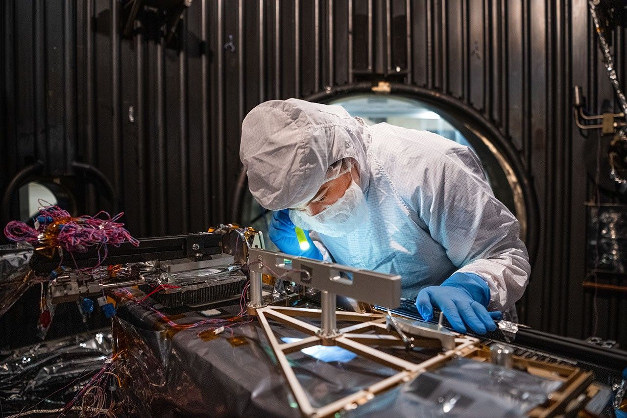 航天图片资料 · A technician in a white clean-room suit and blue gloves uses a small flashlight to inspect a spacecraft instrument inside a dark testing chamber. Colorful wires and metallic components surround the instrument.