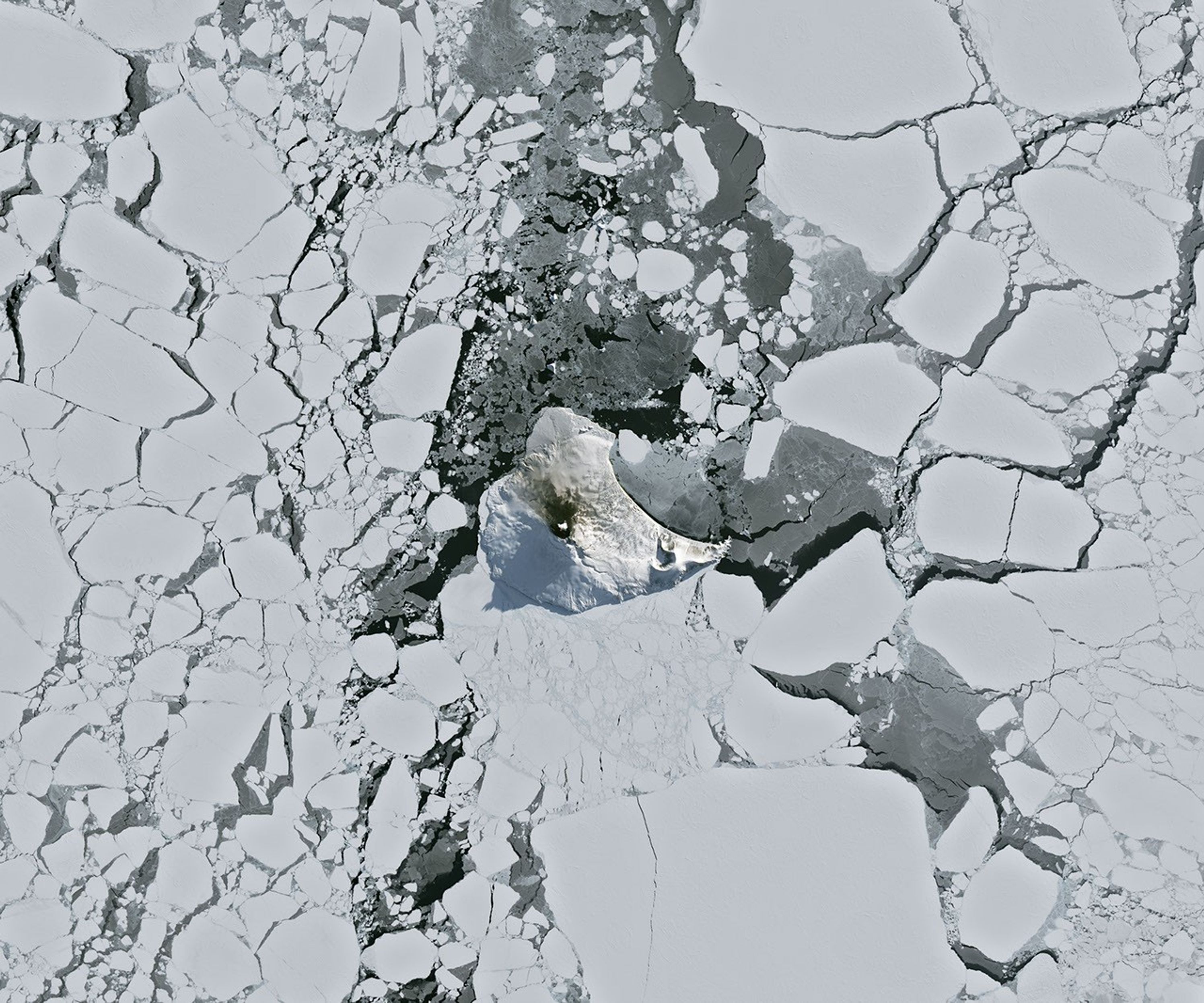 航天图片资料 · A small ice- and snow-covered island with an active volcanic crater at its center is surrounded by drifting pieces of sea ice. A thermal signal and small plume appear in the crater, and ash darkens the snow on the volcano’s northern slopes.