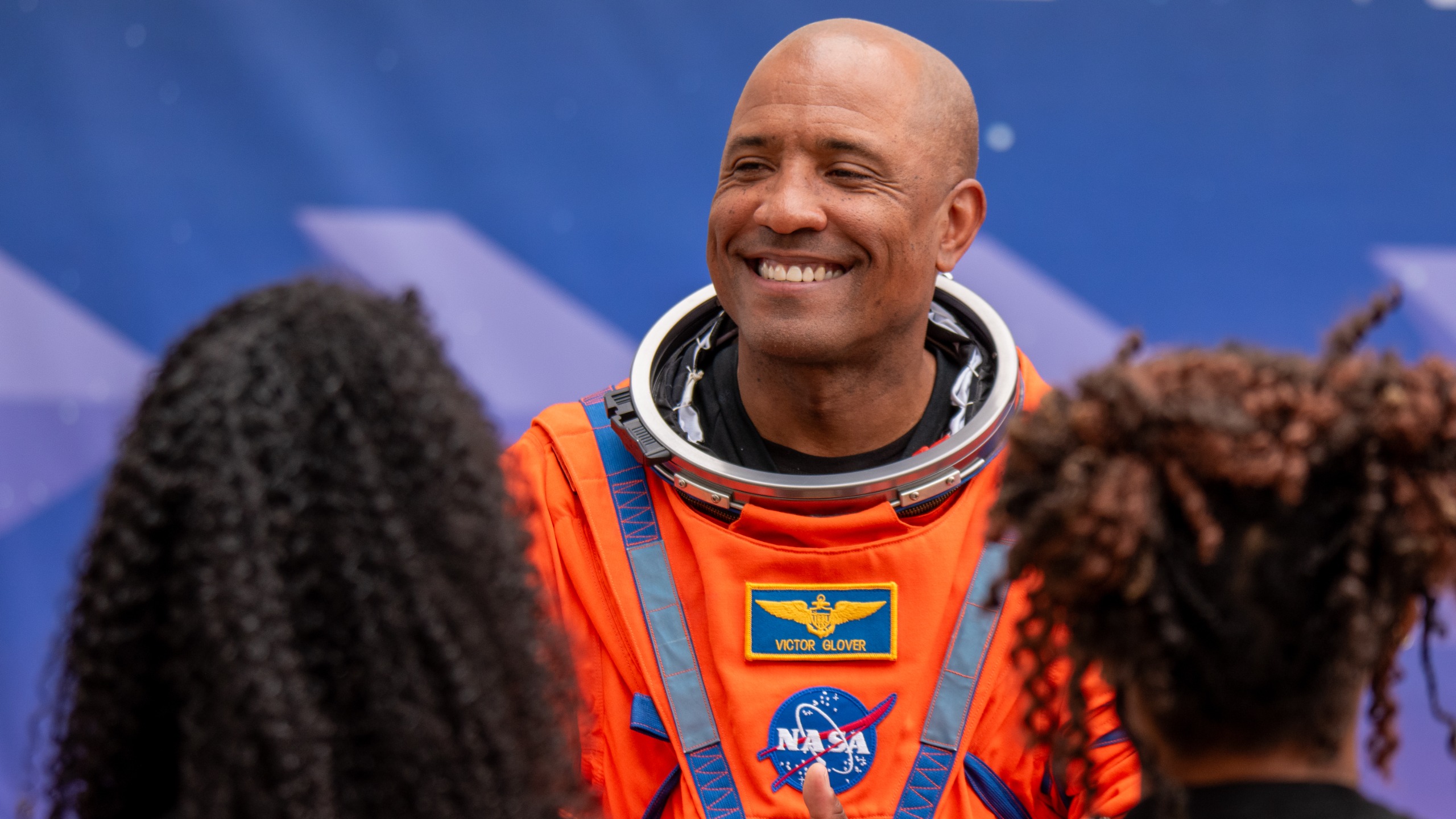 航天图片资料 · A man in an orange space suit smiles at two people, whose back of their heads are unfocused in the forground on left and right.